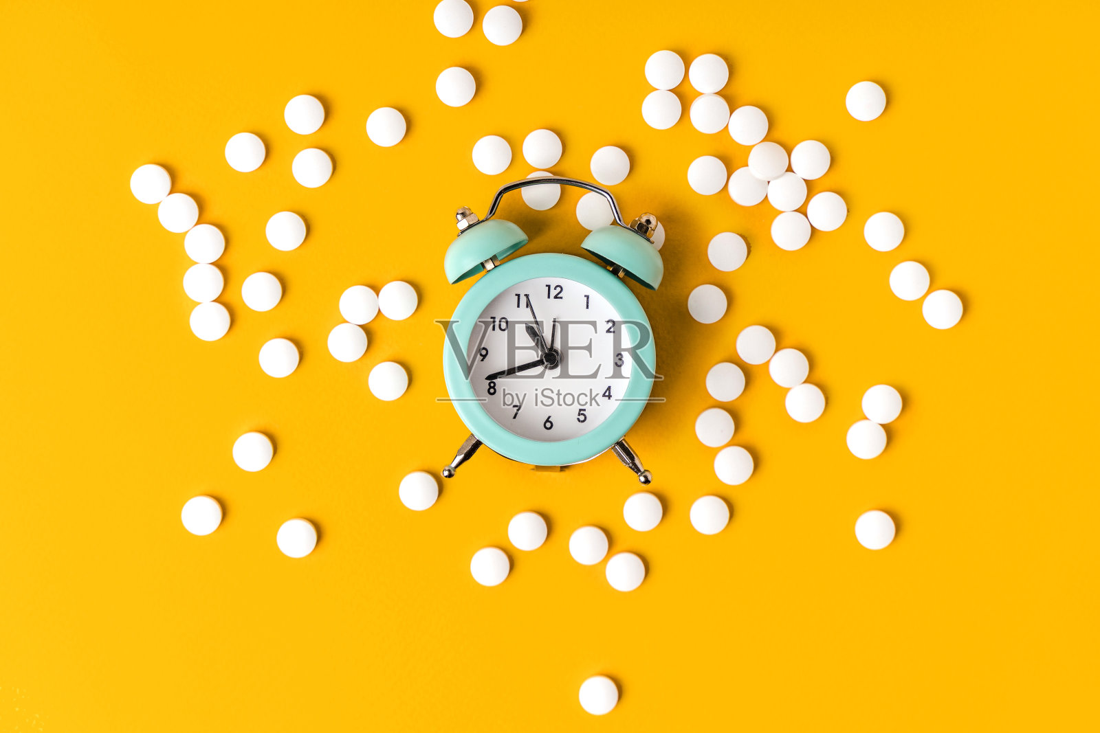 闹钟躺在黄色的背景上，在散落的药丸中间。计划治疗的概念照片摄影图片