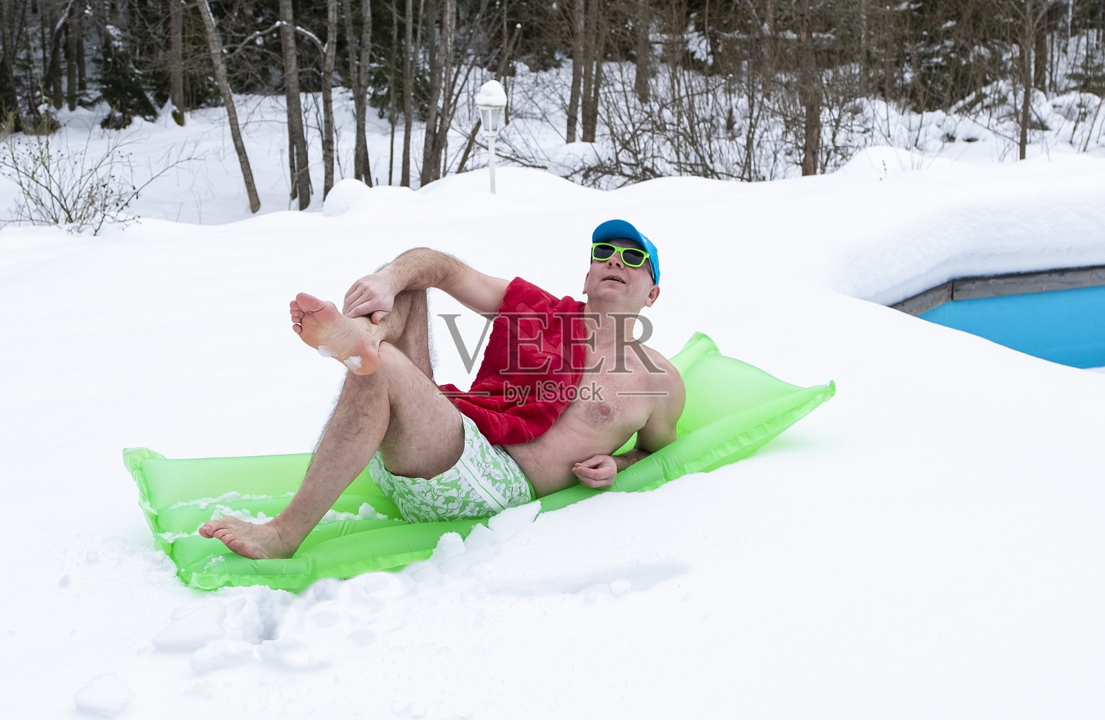 一个男人穿着泳裤，戴着墨镜，在一个充气床垫上结冰。冬天，一个男人在房子后院里的池塘边的雪地里休息。照片摄影图片
