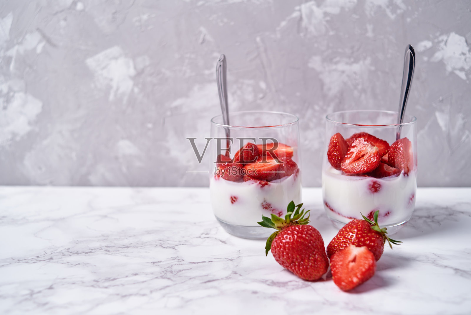 两杯健康的酸奶，新鲜的草莓片和勺子放在白色大理石桌子的背景，复制空间。健康早餐，食物理念照片摄影图片