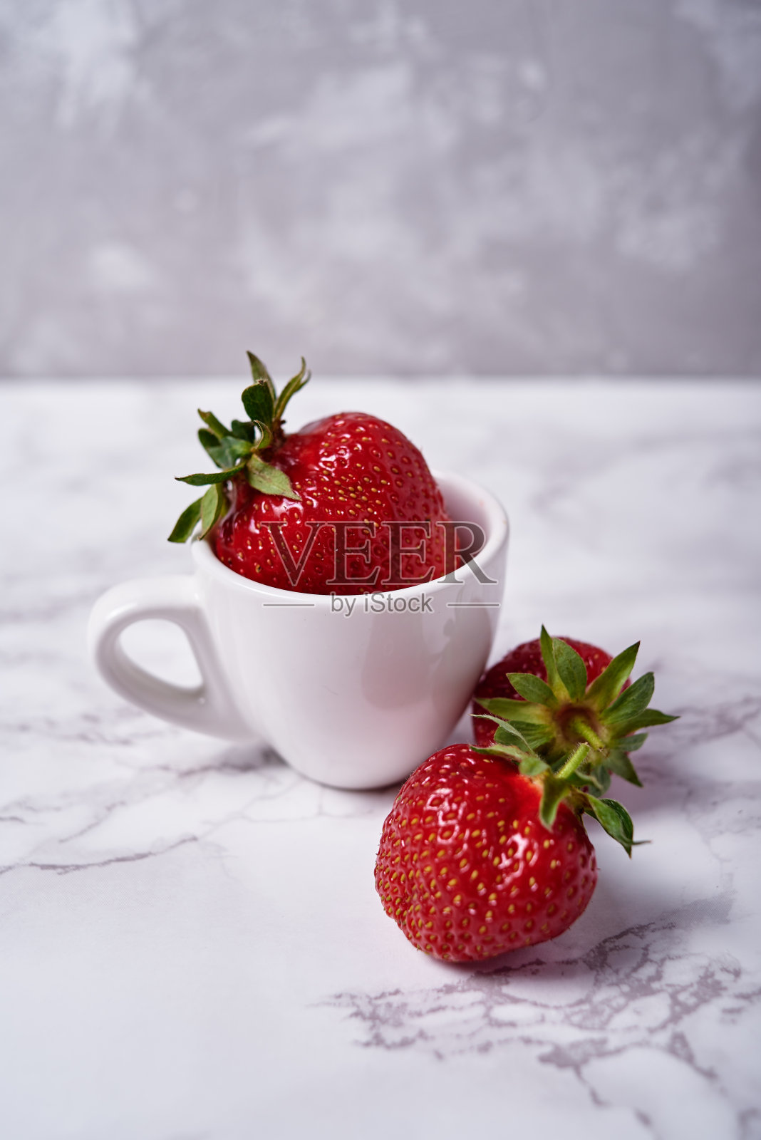 一个大的有机草莓在白色陶瓷咖啡杯在灰色大理石背景，复制空间。健康食品理念，创意静物，特写照片摄影图片