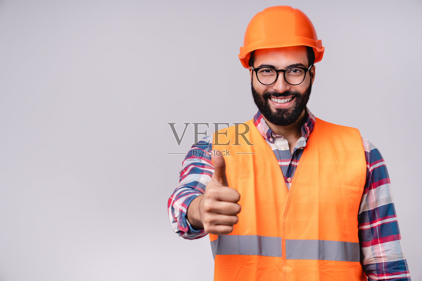 微笑的年轻中东男子显示拇指在安全帽和特殊制服孤立在灰色背景照片摄影图片