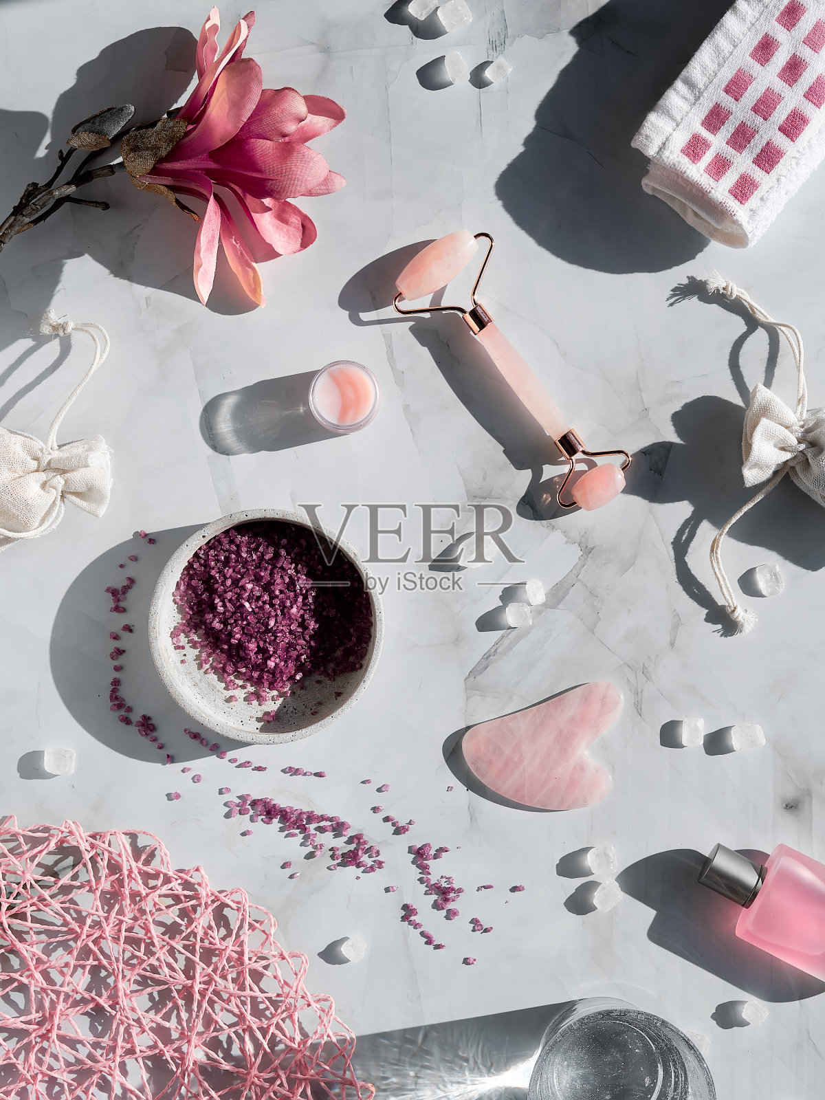 水晶玫瑰石英粉面部按摩辊和刮痧石，平躺在玉兰花大理石桌上。长长的影子。精油瓶子照片摄影图片