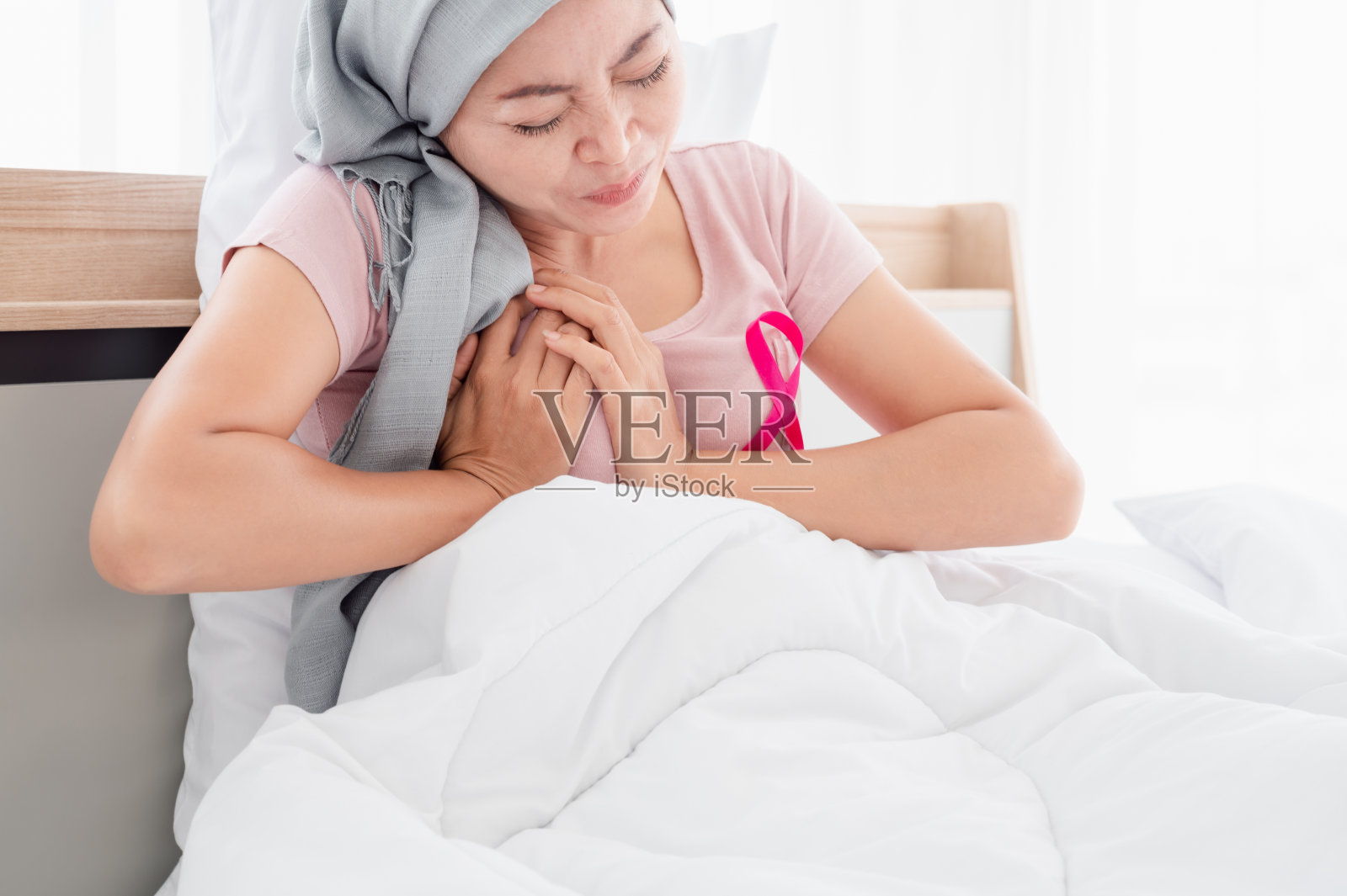 年轻的亚洲妇女戴着头巾坐在床上时由于疼痛双手放在胸前。表示从乳腺癌中恢复乳腺癌概念，癌症预防概念。照片摄影图片