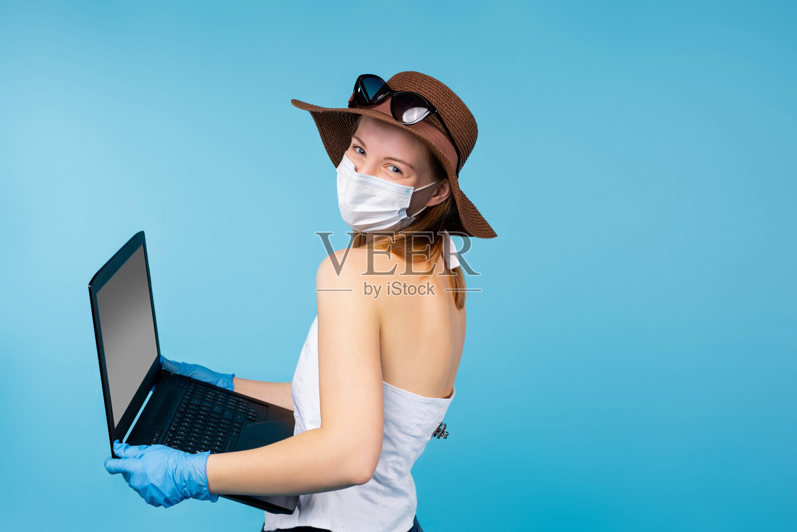 一个白种女孩穿着夏天的衣服，戴着一顶帽子，戴着医用口罩和手套，带着打开的笔记本电脑看着摄像机。大流行期间的旅游和工作概念。照片摄影图片