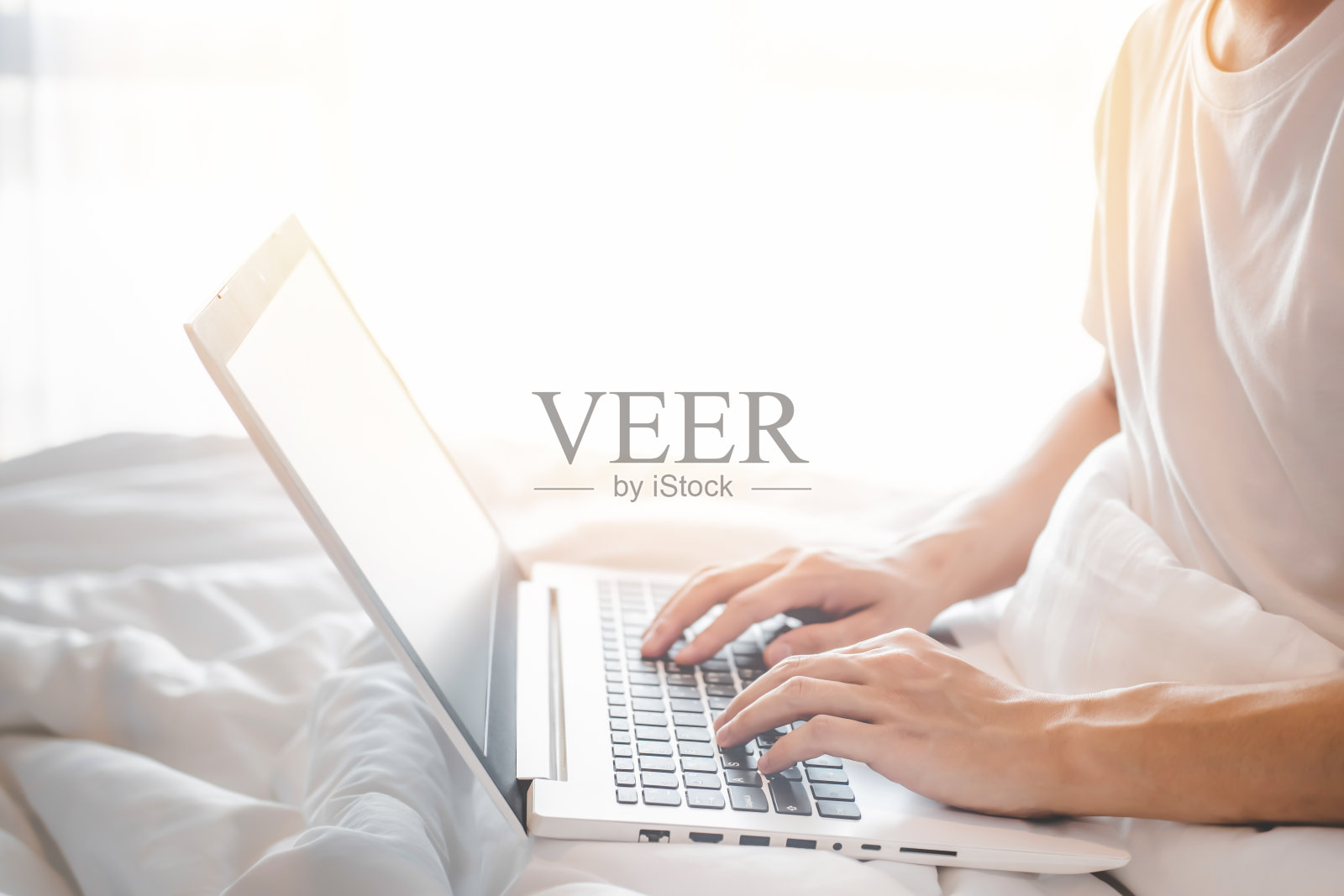 年轻男子的特写穿着白色短袖t恤使用笔记本电脑，坐在白色的床和白色的毯子与阳光。在家工作的概念，学生在家里的卧室在线学习照片摄影图片