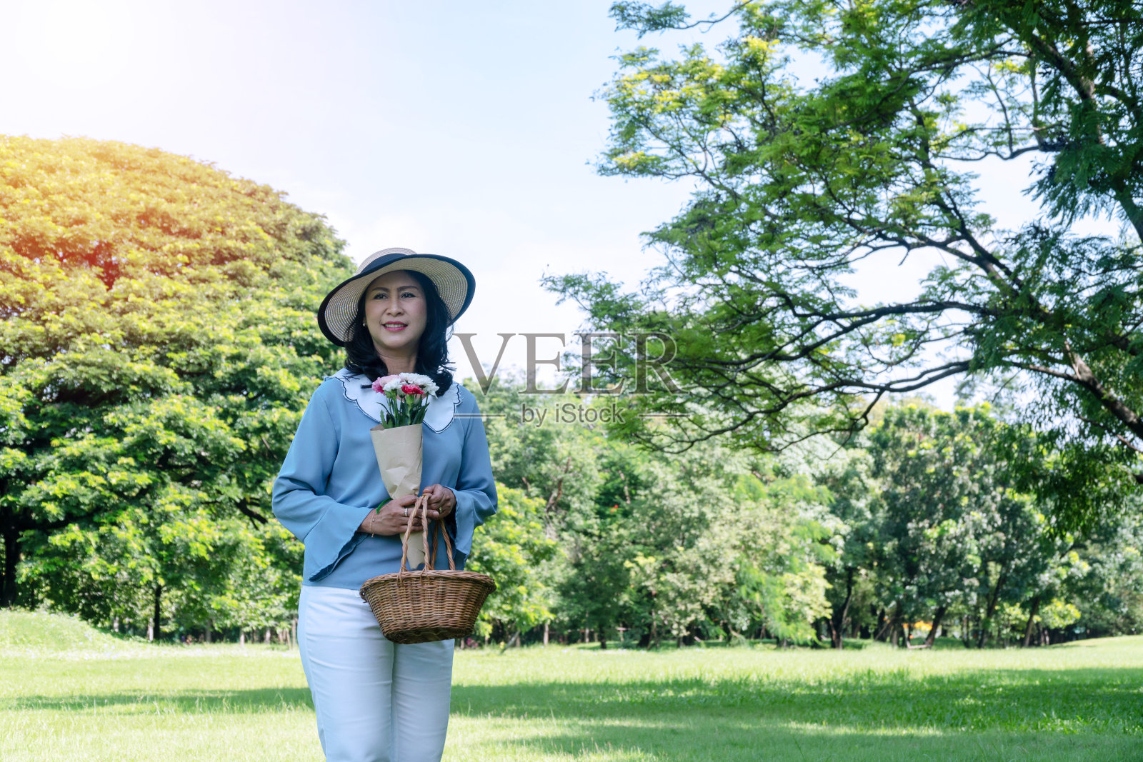 老年人生活方式的概念。欢快微笑的50多岁的老年妇女，戴着帽子，手里拿着水果篮和一束花，看着公园里的相机。照片摄影图片