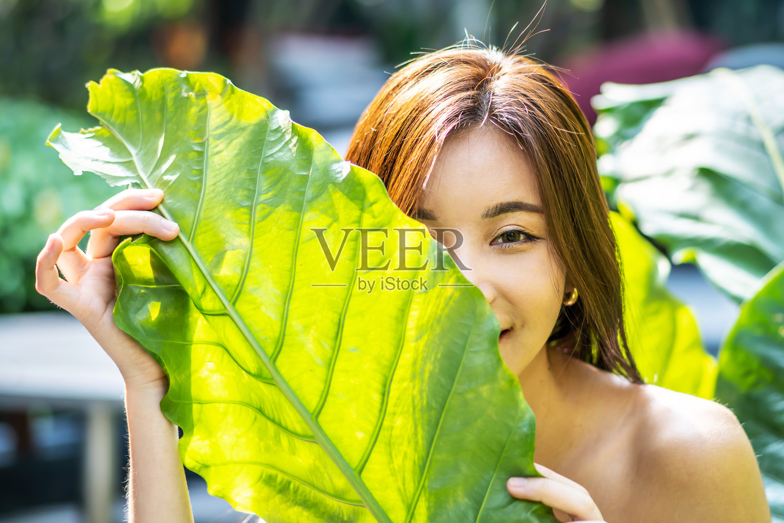 一个美丽的亚洲年轻女子的肖像与自然的化妆持有一个大绿叶在模糊的绿色背景。水疗和健康。青春、青少年护肤理念照片摄影图片