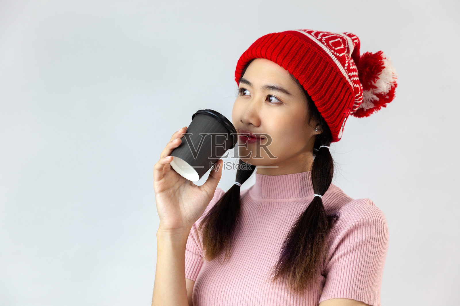 一个年轻的亚洲女人戴着红色的羊毛帽和粉红色的t恤拿着一个咖啡纸杯在白色的背景照片摄影图片