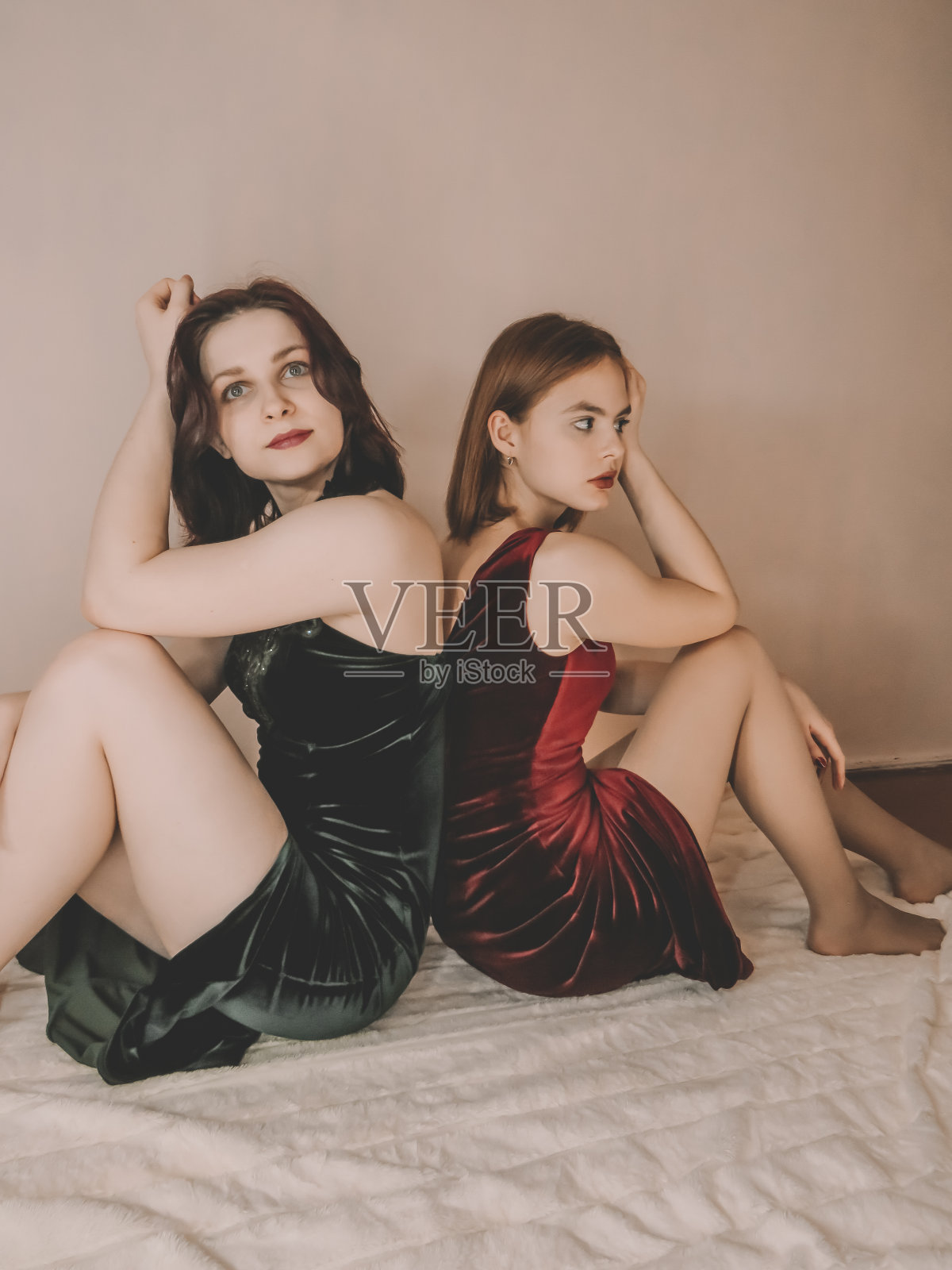 两个苗条迷人的年轻女子背靠背坐在米色毯子上，靠在一面墙上，一个是黑发，一个是棕色头发，穿着翠绿色和紫红色的天鹅绒晚礼服，画着明亮的晚妆，修着鲜红色的指甲照片摄影图片