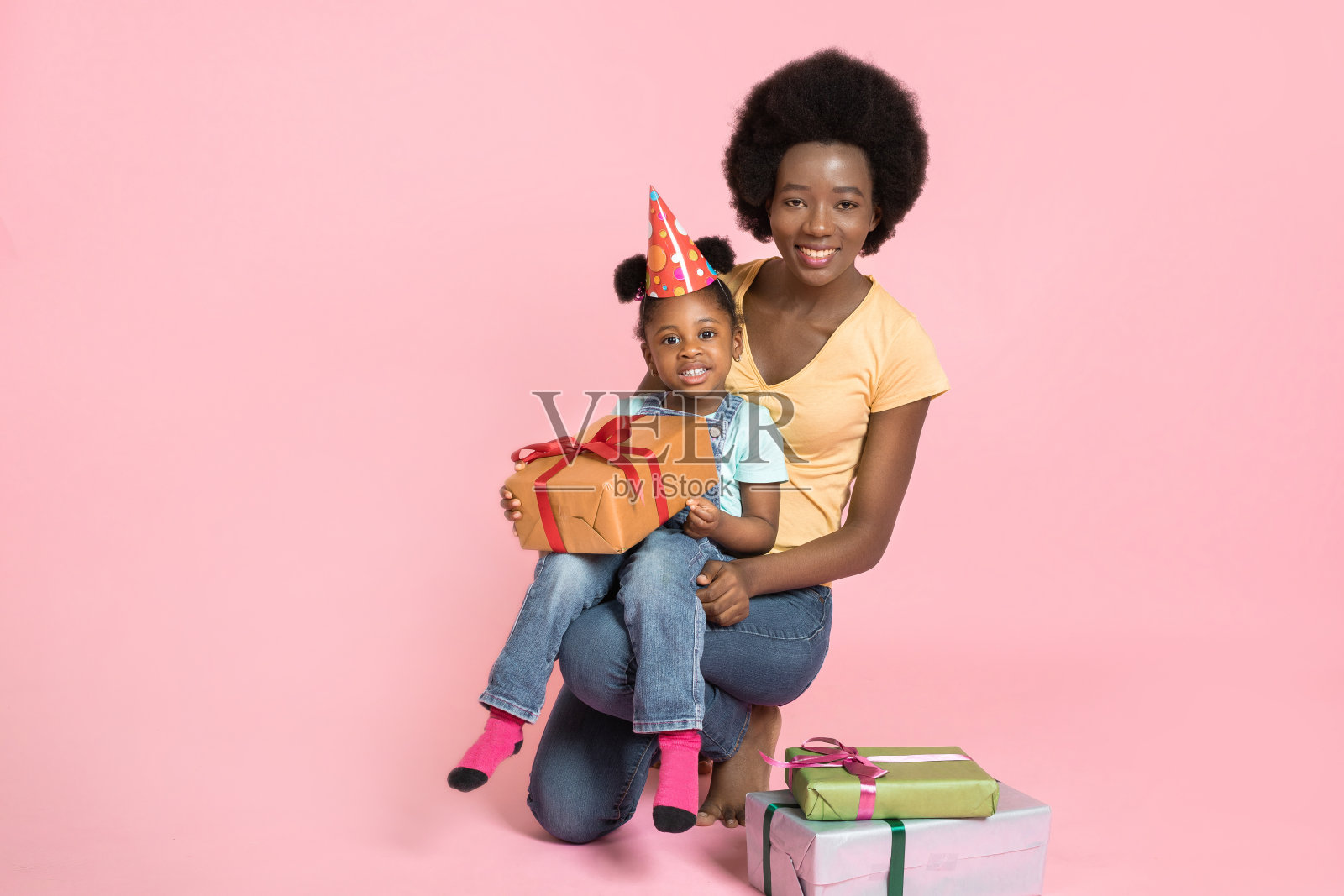 孩子的生日、节日庆祝概念。可爱快乐的小非洲女孩在派对帽子，拿着黄色的礼物盒，而坐在她妈妈的腿上，在粉红色的工作室背景照片摄影图片