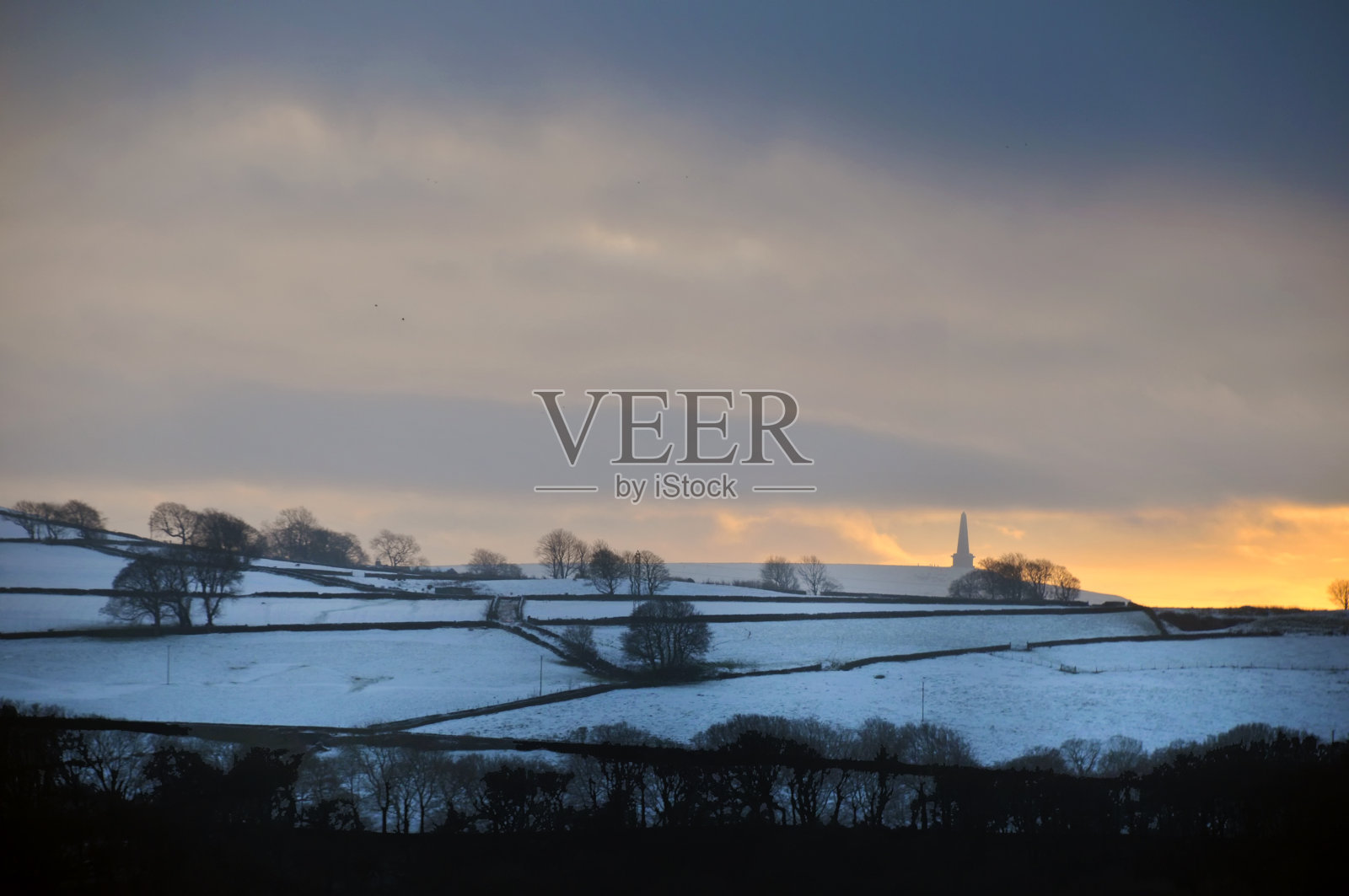 在一个多云的黄昏的冬天天空中，闪烁着橙色的日落，雪地覆盖着田野，远处是西约克郡卡尔德代尔的斯塔德利狗鱼纪念碑照片摄影图片