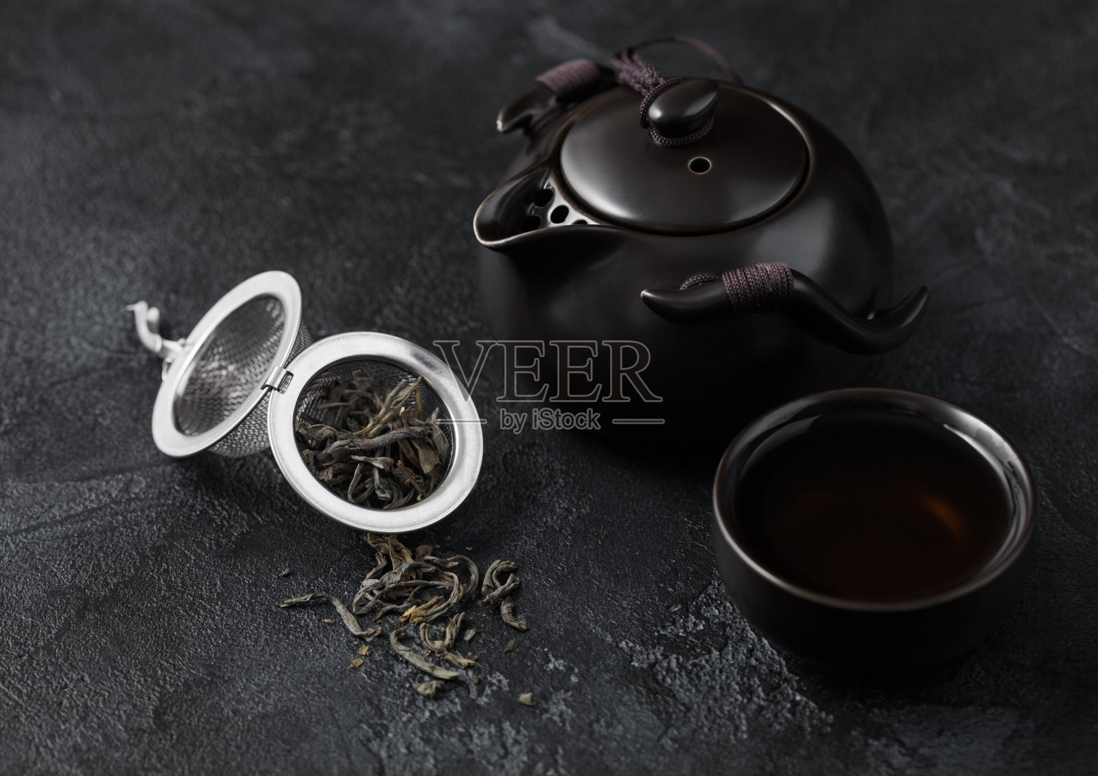 松散的绿色有机茶在过滤灌浆器和陶瓷茶壶和杯子上深色。照片摄影图片