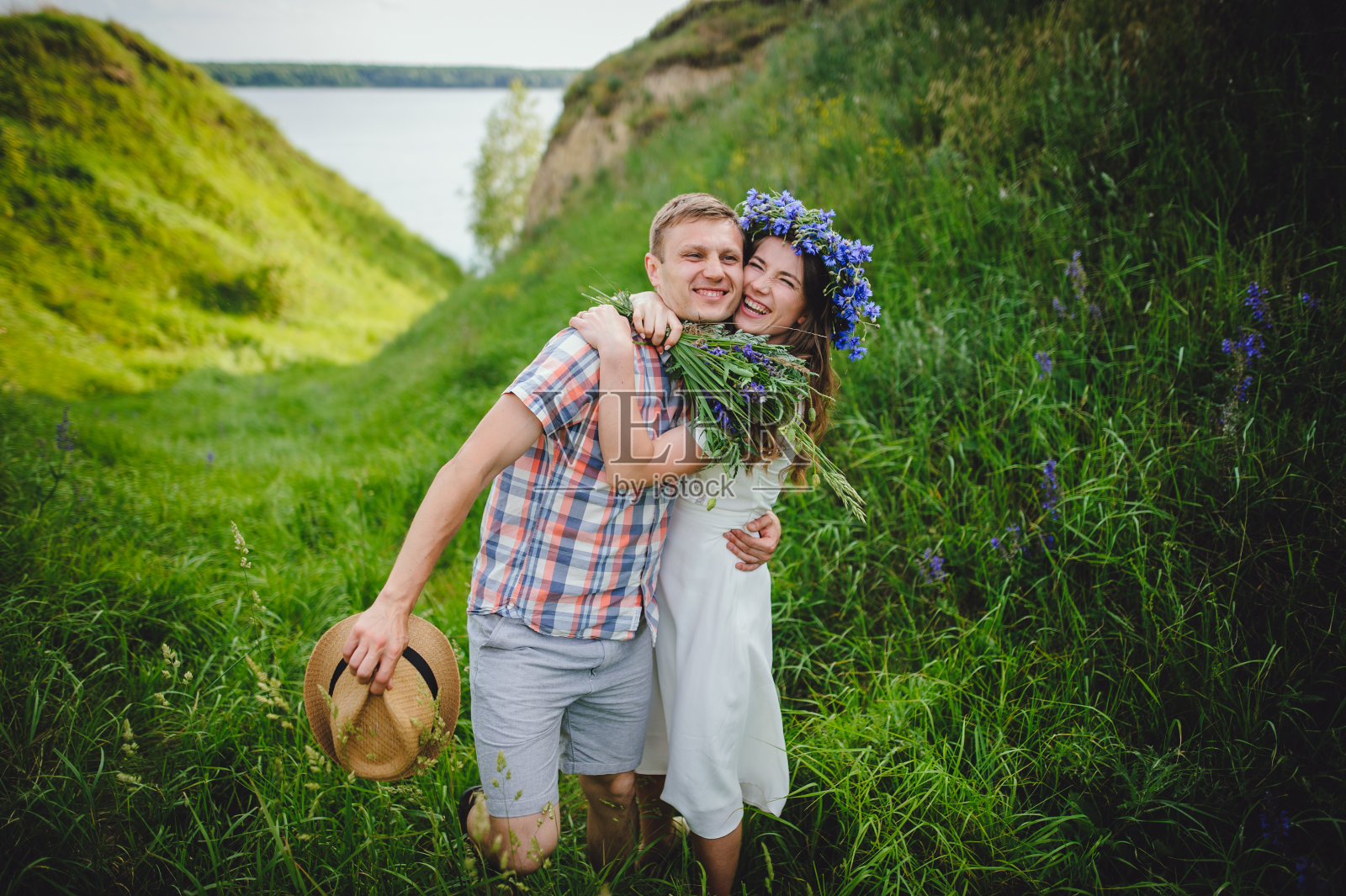快乐的女孩抱着她的男朋友，手里拿着一束野花。有趣的情绪。美丽的女孩在花圈和时尚的男孩花时间在户外在一个阳光明媚的日子肖像。爱情故事。照片摄影图片