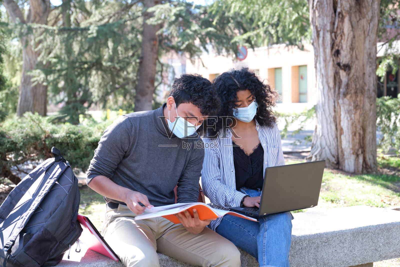 两个戴着防护面罩的拉丁学生一起坐在户外的长椅上学习。大学校园的新常态。照片摄影图片