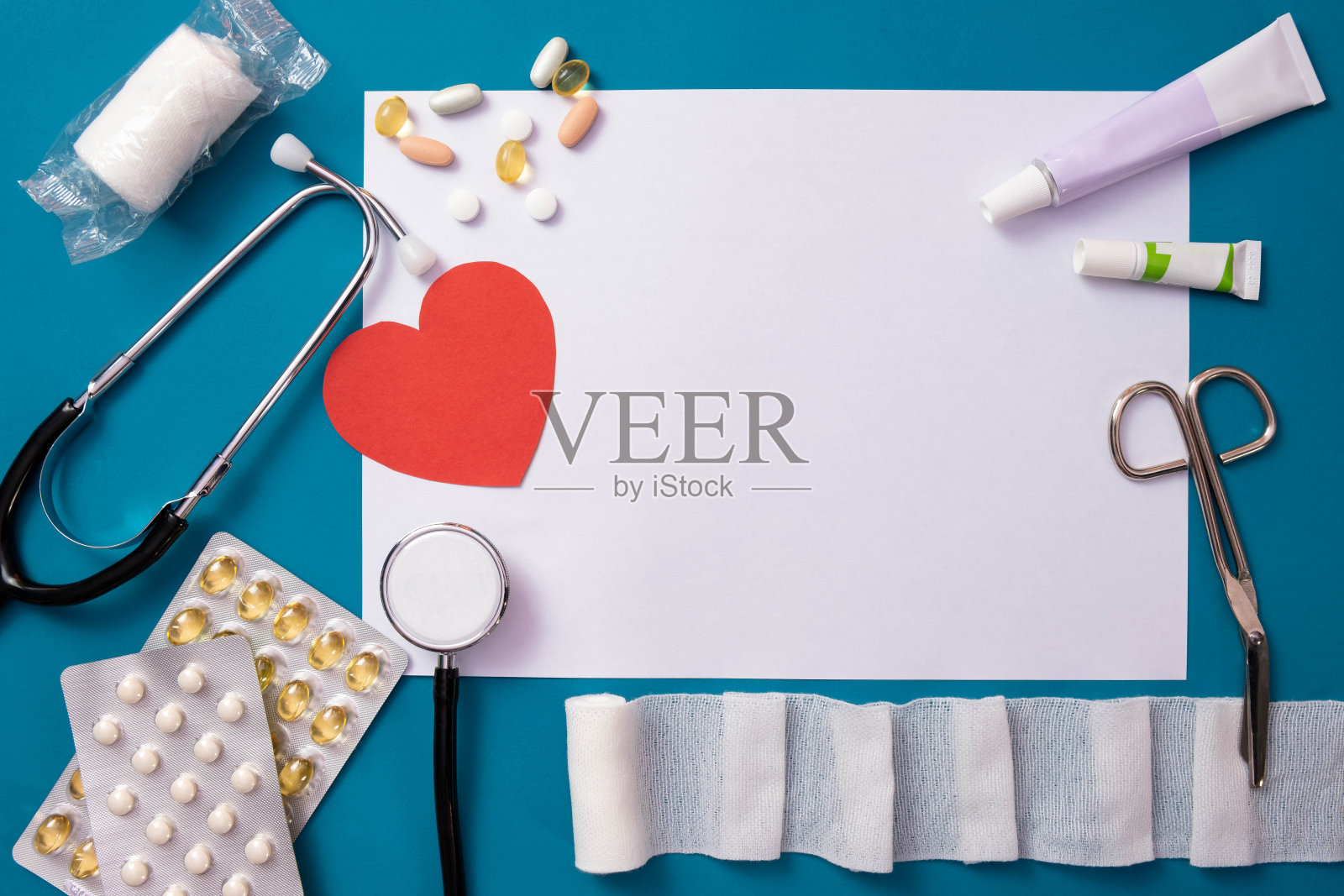 桌上放着绷带、剪刀、红心、听诊器、药丸和一张白纸，背景是蓝色的照片摄影图片