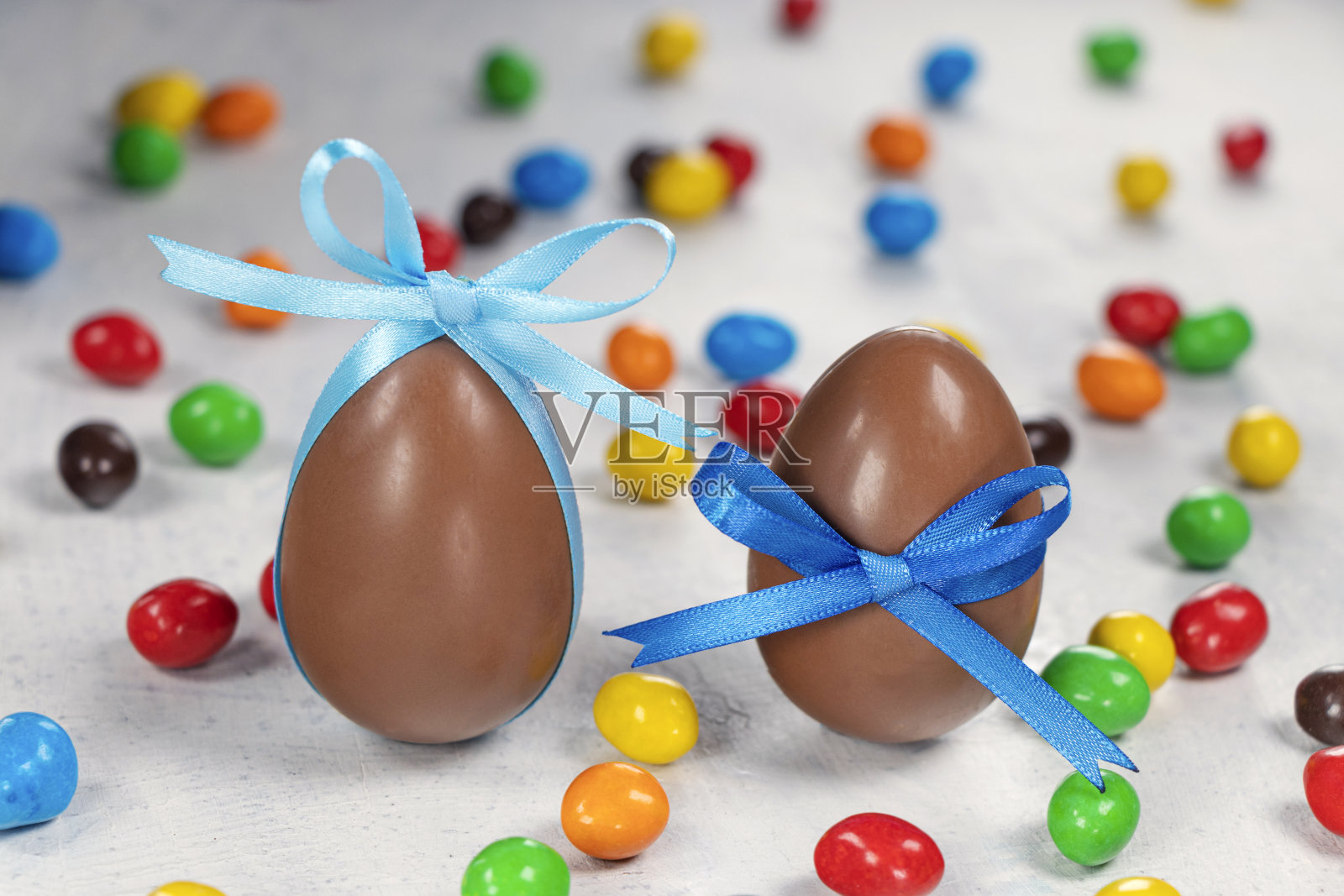 两个大的巧克力彩蛋，上面有一个蓝色的蝴蝶结，白色的混凝土背景上还有五颜六色的小彩蛋照片摄影图片