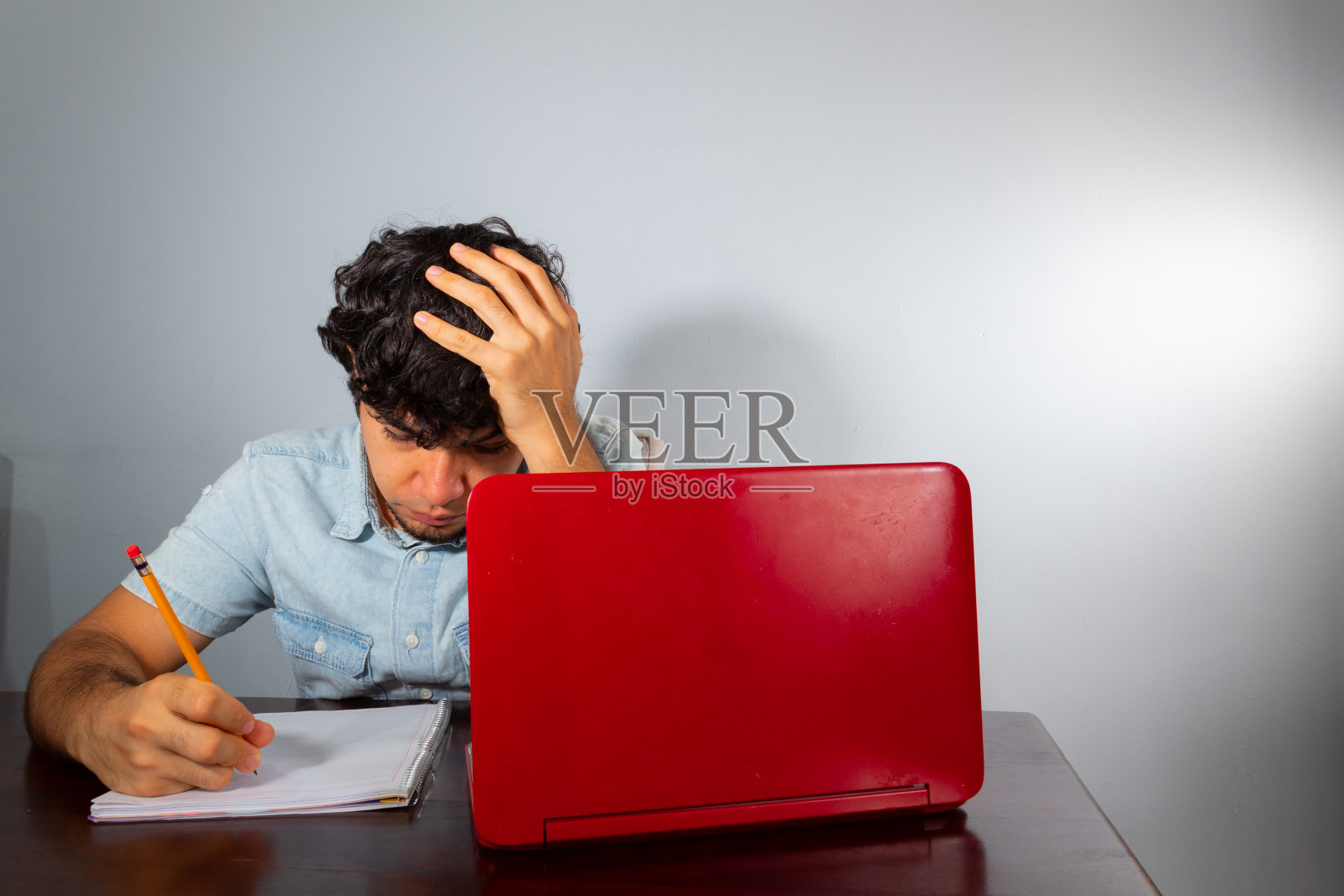 年轻的西班牙卷发男子在家学习，用笔记本电脑，用铅笔在笔记本上写作照片摄影图片