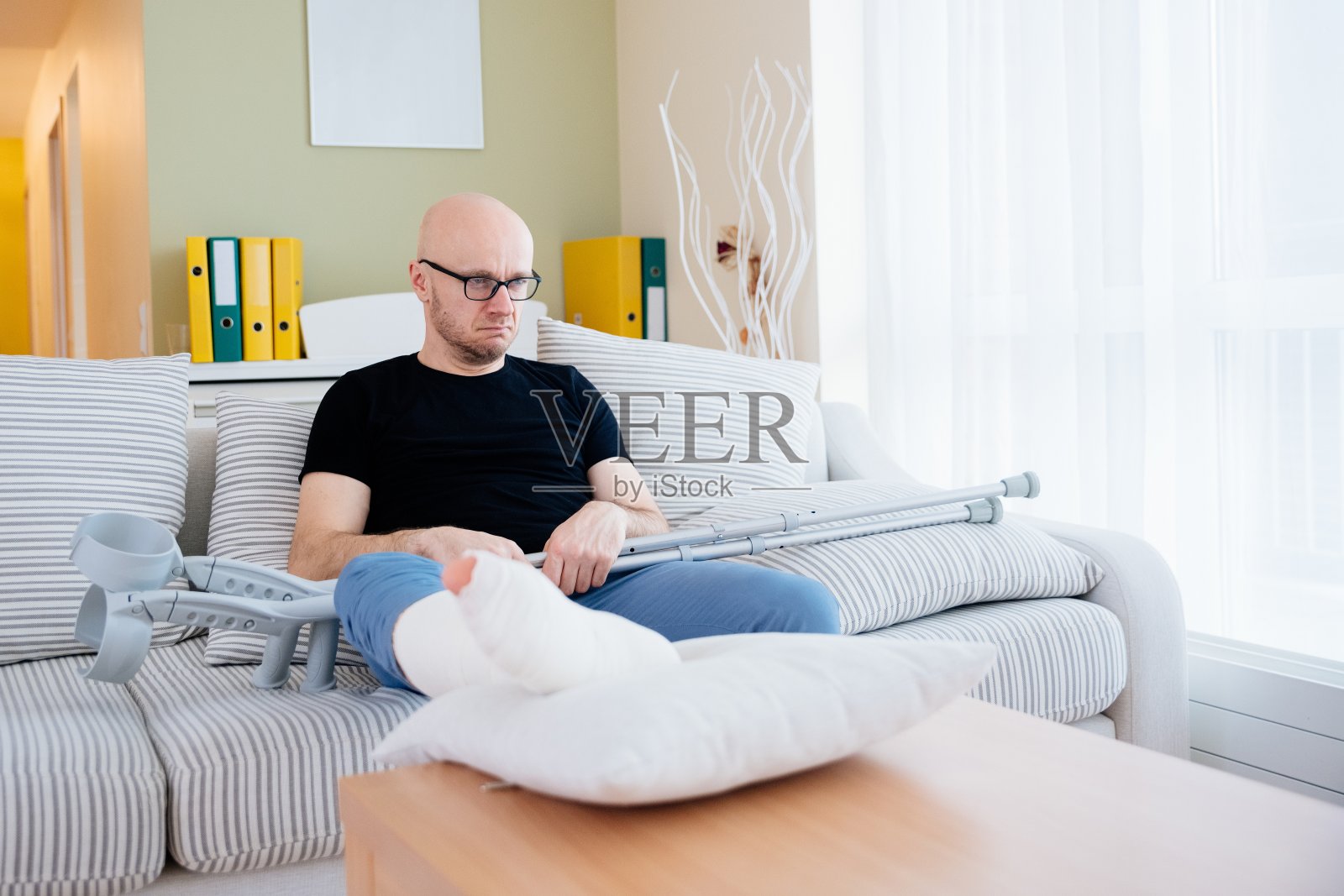 悲伤，愤怒的男人坐在家里的沙发事故后。他手里拿着拐杖。照片摄影图片