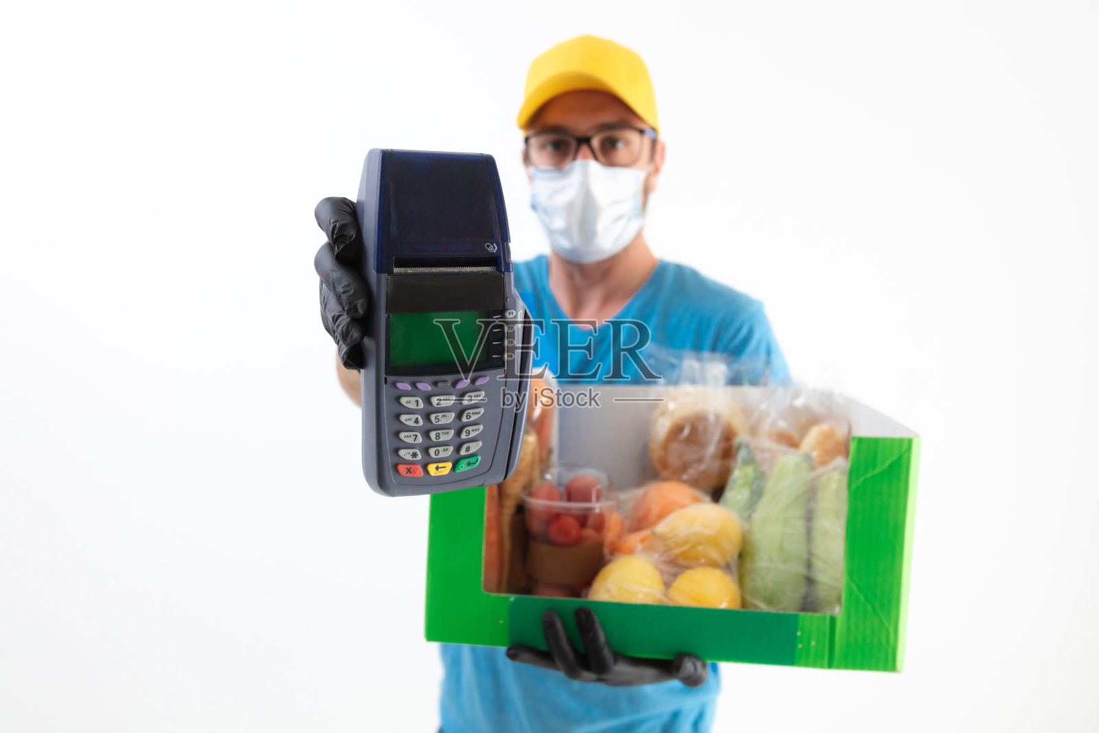 一个戴着防护面具站在白墙前的快递员，手里拿着一个装着杂货和非接触式支付POS机的盒子。照片摄影图片