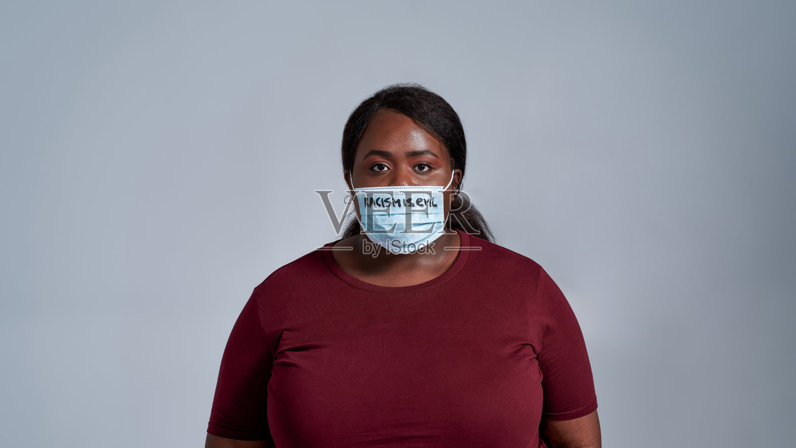 严肃的加码年轻非洲裔美国妇女的肖像，穿着休闲衣服戴着带有种族主义的防护面罩，是邪恶的文字看着相机照片摄影图片