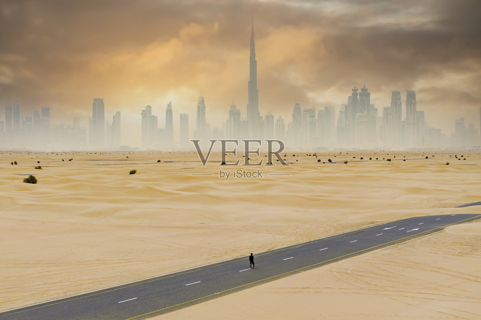 航拍视角：迪拜天际线背景下，行人漫步于沙丘覆盖的荒凉道路照片摄影图片