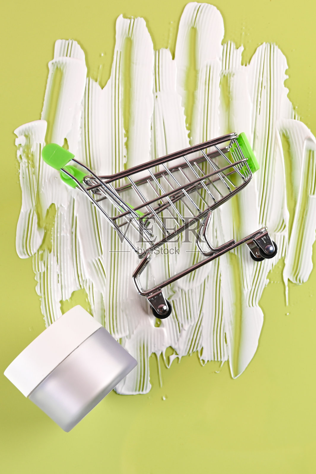 俯视图白色化妆品罐与奶油和购物手推车在一个绿色的背景。在线家庭购物照片摄影图片