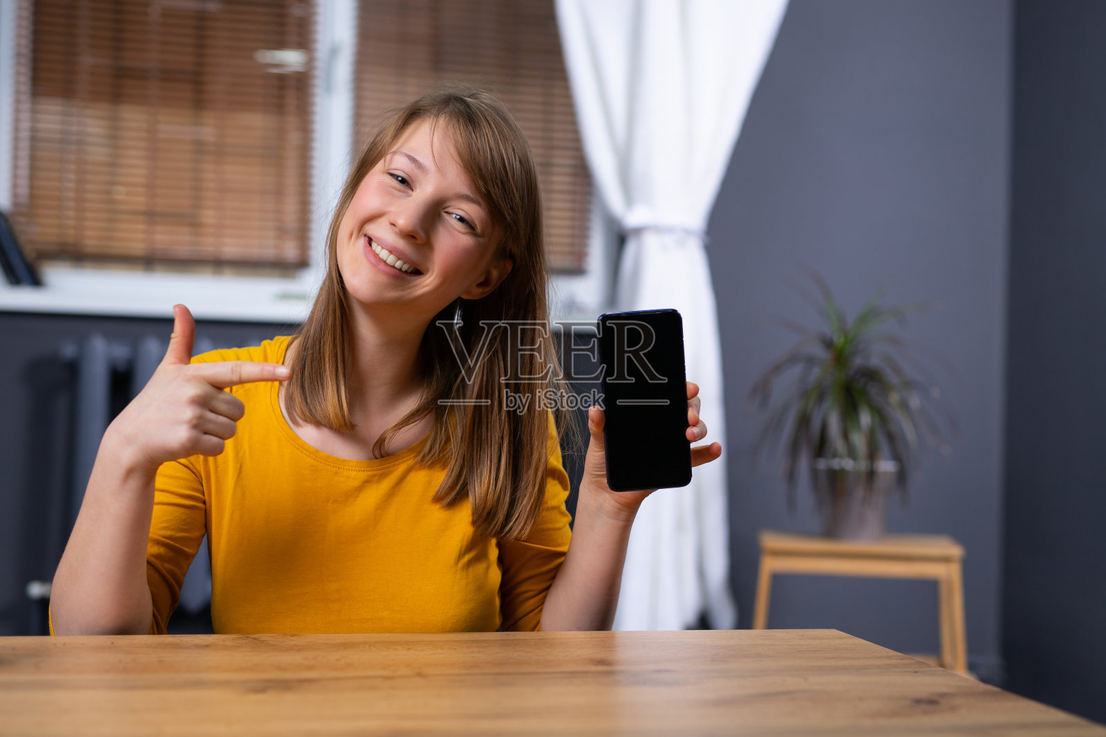 金发女人在家里办公室，微笑着，拿着现代智能手机的空白模拟屏幕，十几岁的女性用她的食指指向它。空白的广告或促销文字。照片摄影图片