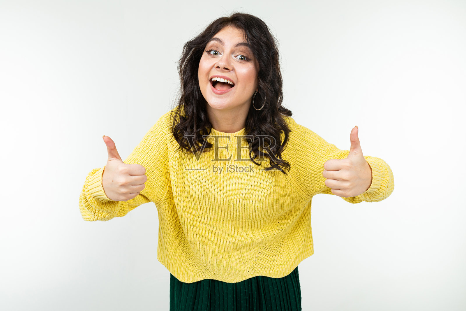 微笑的黑发女孩穿着黄色的毛衣和绿色的裙子显示拇指在白色的背景照片摄影图片