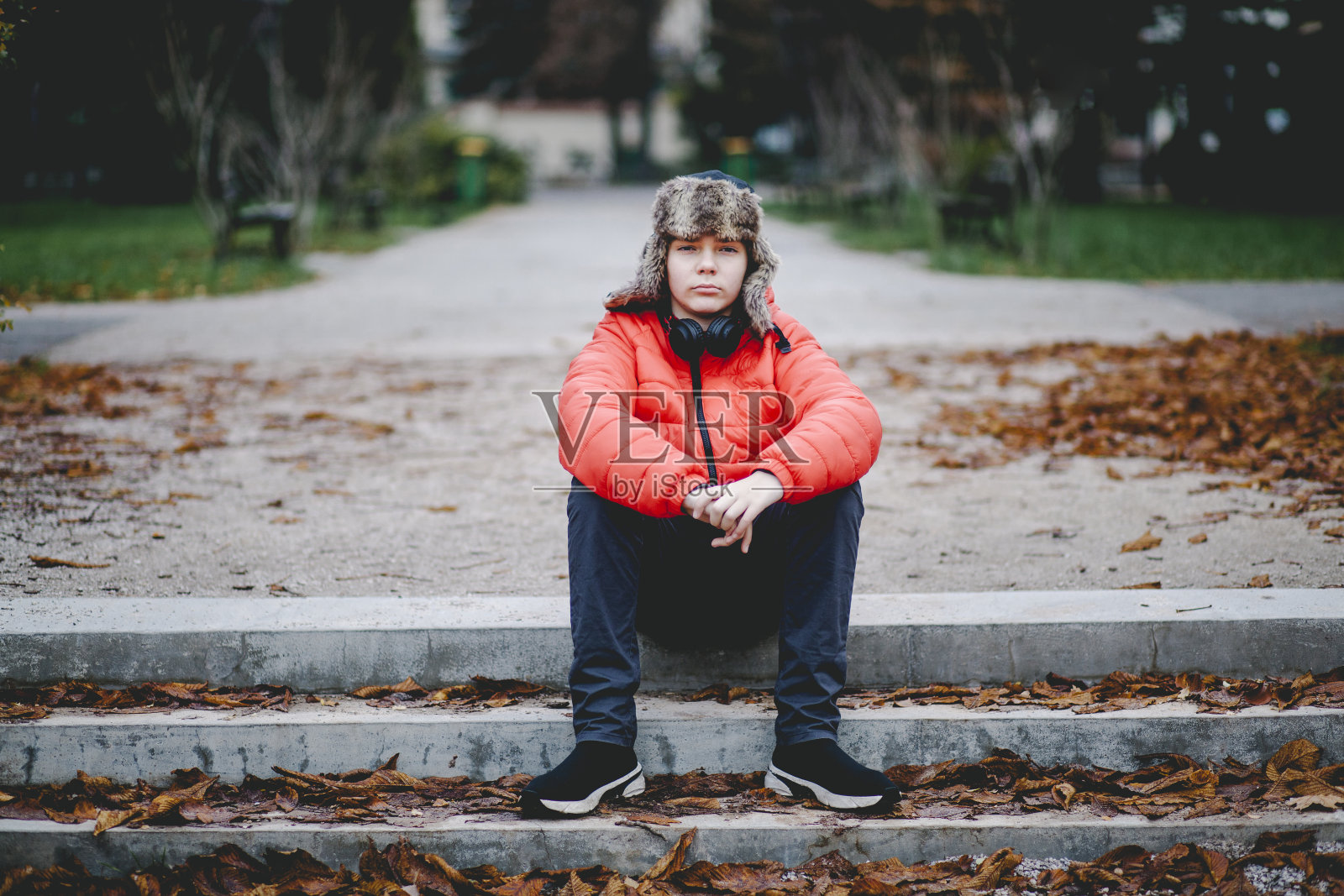 严肃可爱的十岁男孩穿着红色的暖夹克，头戴帽子坐在公园的台阶上，双手合十放在膝盖上，看着镜头，等待着，冷静着，盯着照片摄影图片