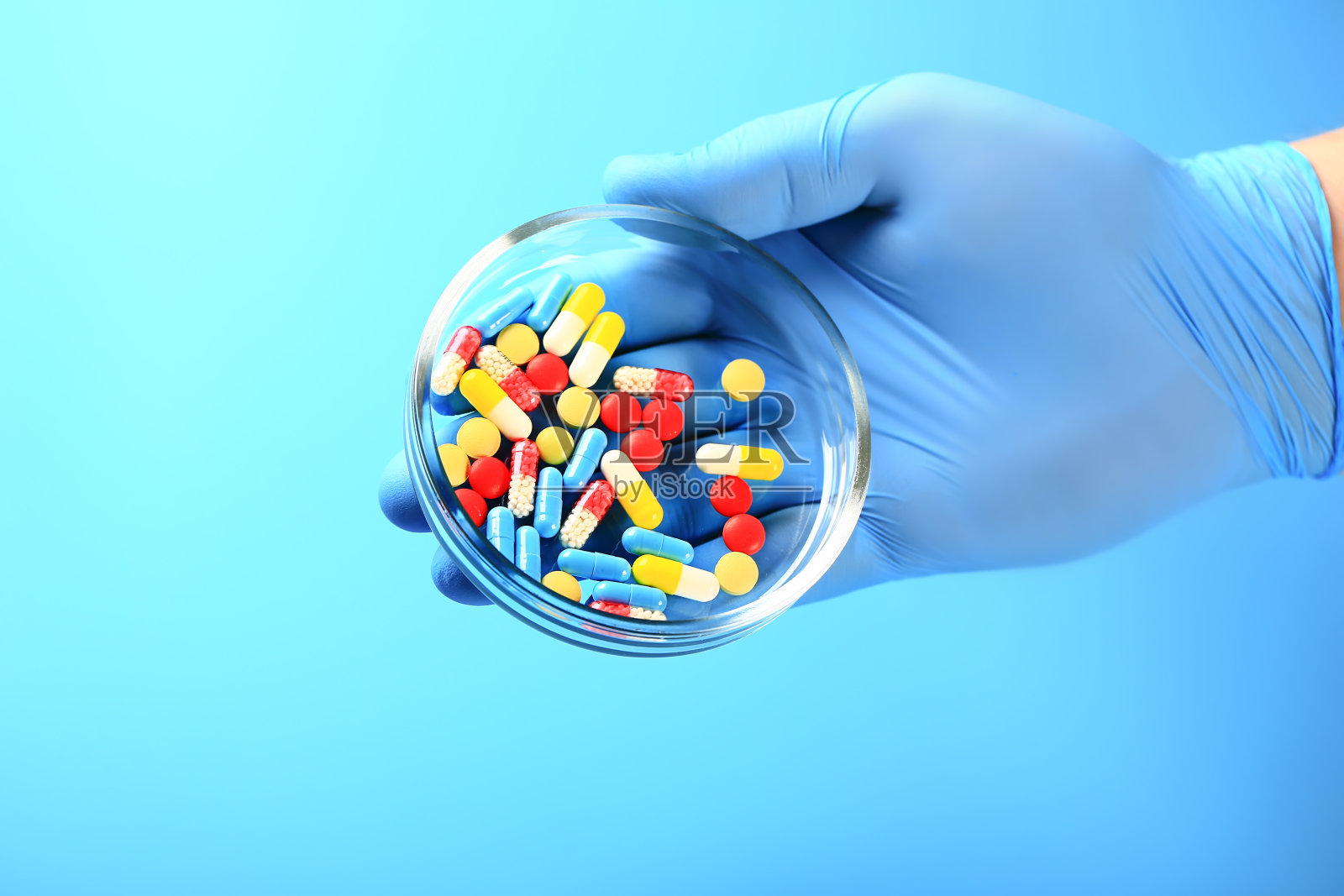 手在一个蓝色的乳胶手套拿着一个玻璃杯与多种颜色的药丸。照片摄影图片