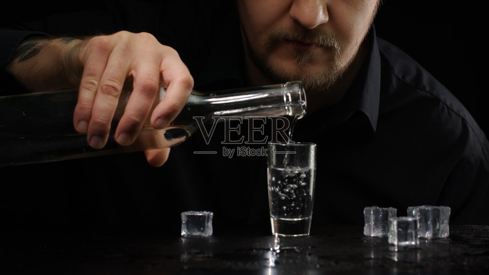 一名酒鬼将冰冻的伏特加从瓶子中倒入装有冰块的小酒杯中，背景为黑色照片摄影图片