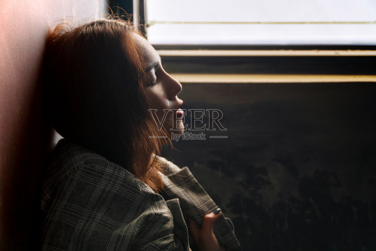 脸色沮丧的年轻女子棕色头发独自站在窗前，同时闭上眼睛，思考着什么。孤独、忧郁、女商人。照片摄影图片