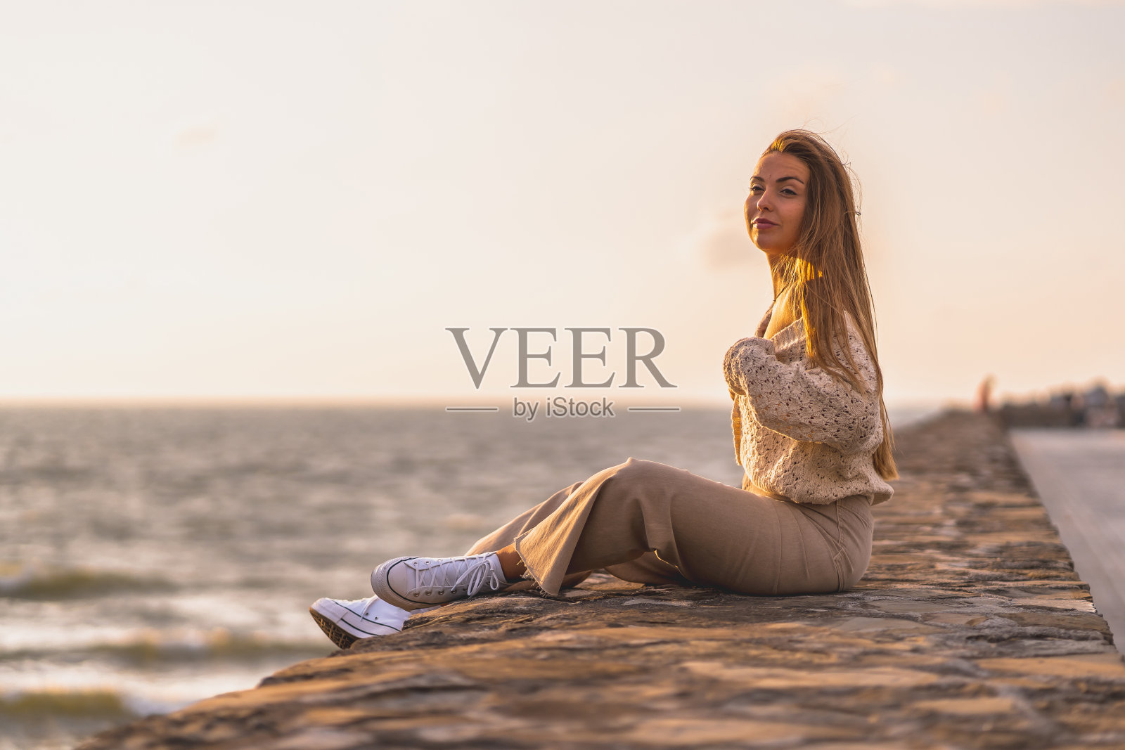 夏天的生活方式，一个年轻的金发白种女人坐在海边，穿着白色露脐上衣和灯芯绒裤子。在一个夏天的下午，在我闭着眼睛的黄金时刻照片摄影图片