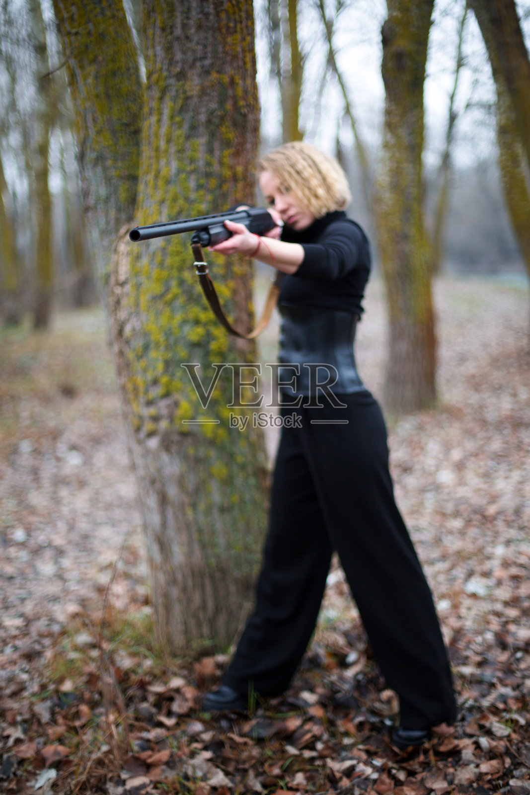 在秋天的森林里，一个穿着黑色套装和紧身胸衣的金发女人在树干前瞄准了一把猎枪照片摄影图片