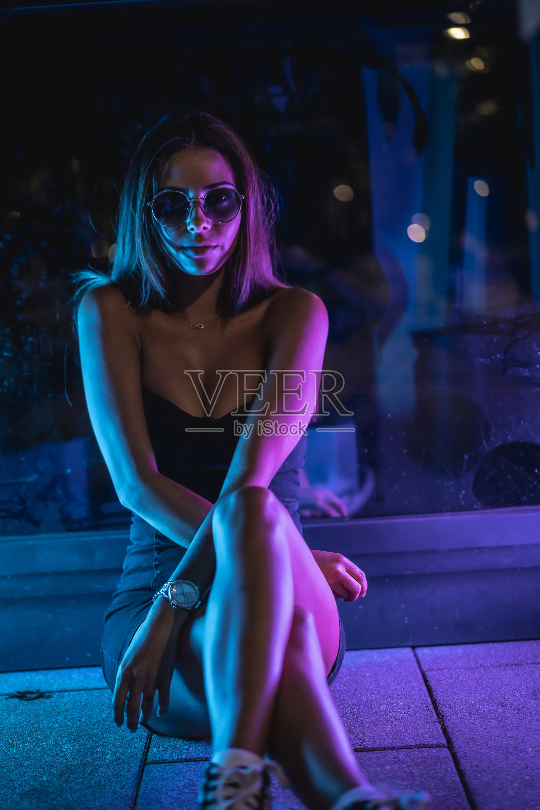 一位身穿黑色连衣裙的年轻白人女性，在紫色和蓝色LED灯反射的黑色水晶的照射下，静静地坐在地板上。夜晚城市摄影照片摄影图片