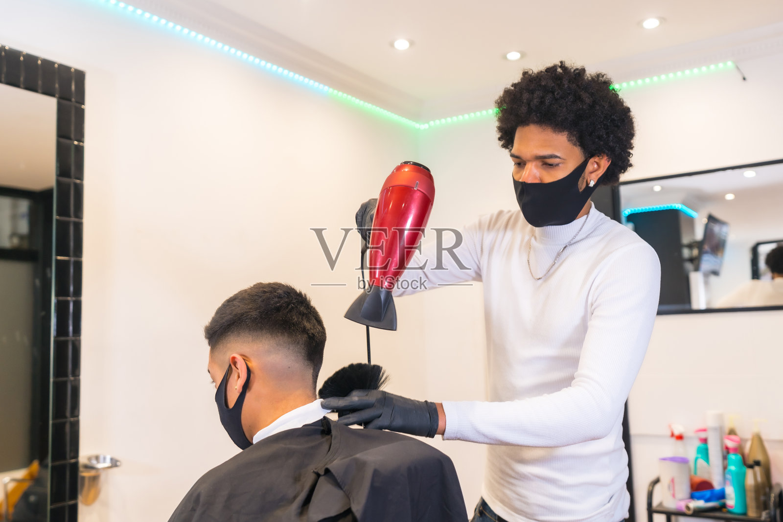 年轻的美发师戴着口罩和手套，用吹风机吹干客户的头发。新冠肺炎大流行期间美发业的安全措施照片摄影图片