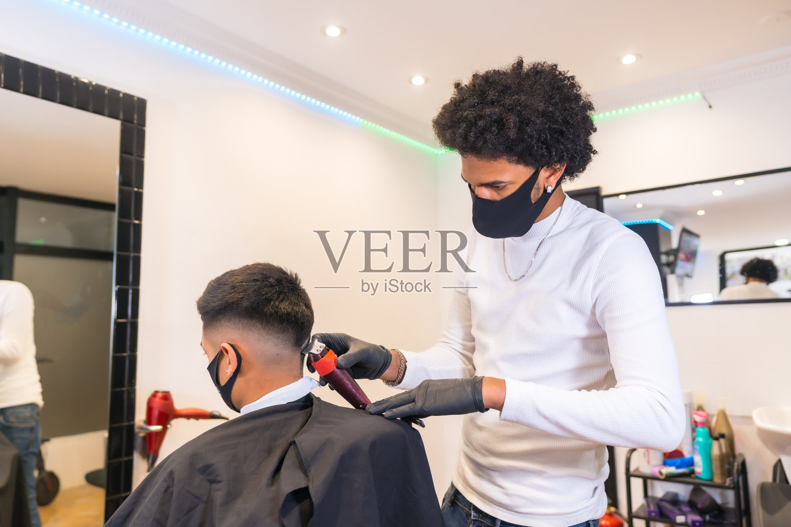 年轻的理发师戴着面具和手套剪头发。新冠肺炎大流行期间美发业的安全措施照片摄影图片