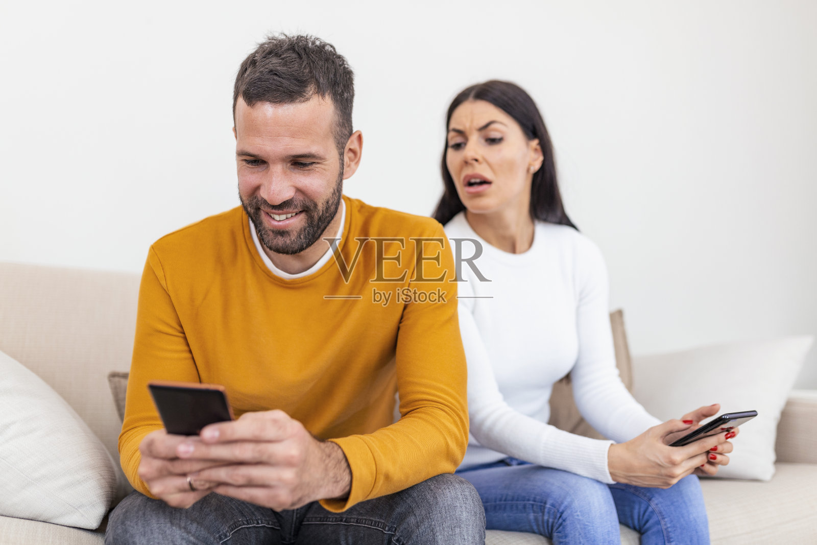 快乐微笑的男人背对着妻子，读着情人发来的手机短信，忧心忡忡的女人坐在他旁边，试图偷看屏幕。欺骗和不忠的概念照片摄影图片