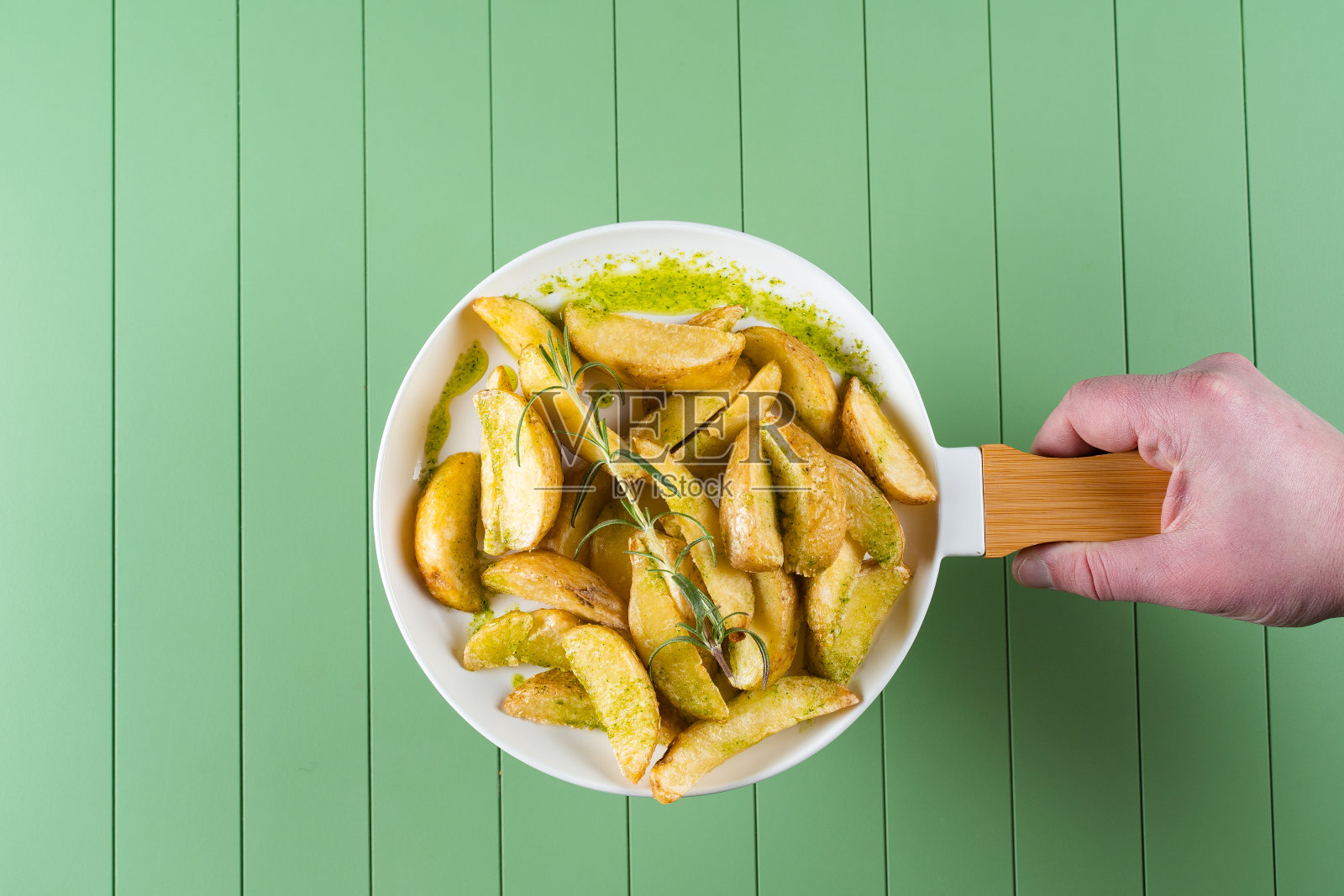 烤土豆配上绿色酱汁和迷迭香，放在煎锅状的白色盘子上。绿色的桌子上放着香蒜酱炸土豆。照片摄影图片