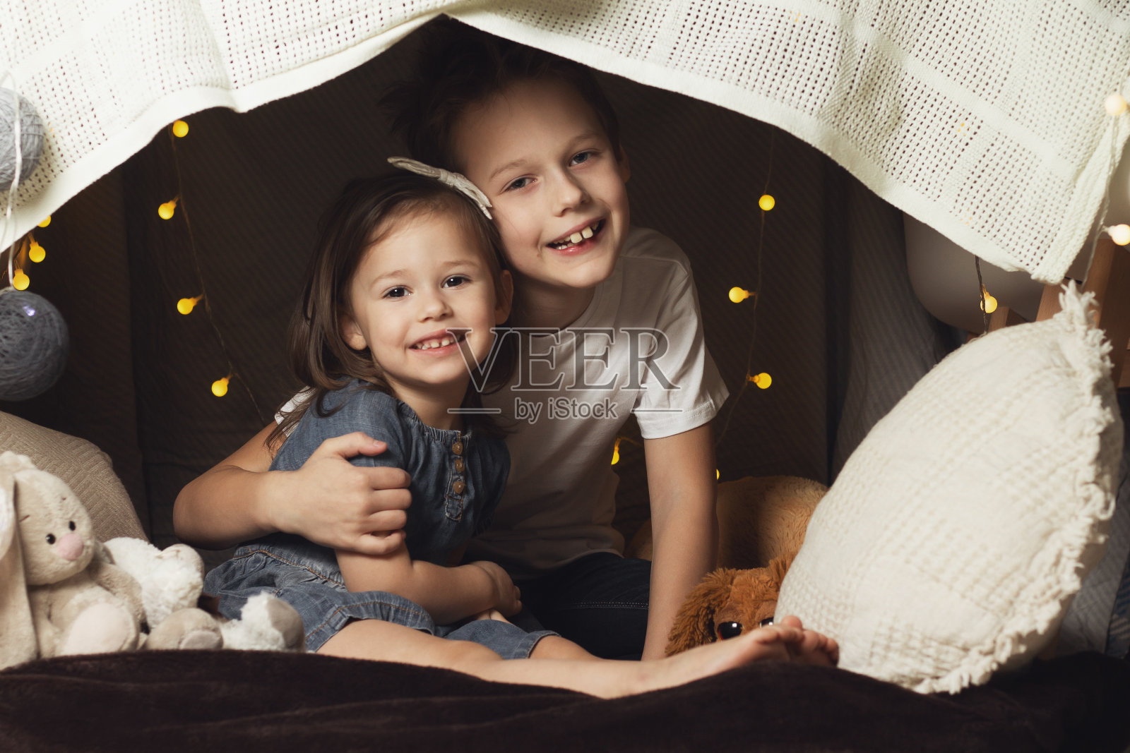 兄弟姐妹们住在用椅子和毯子搭成的小屋里。弟弟和妹妹在家里又笑又玩照片摄影图片