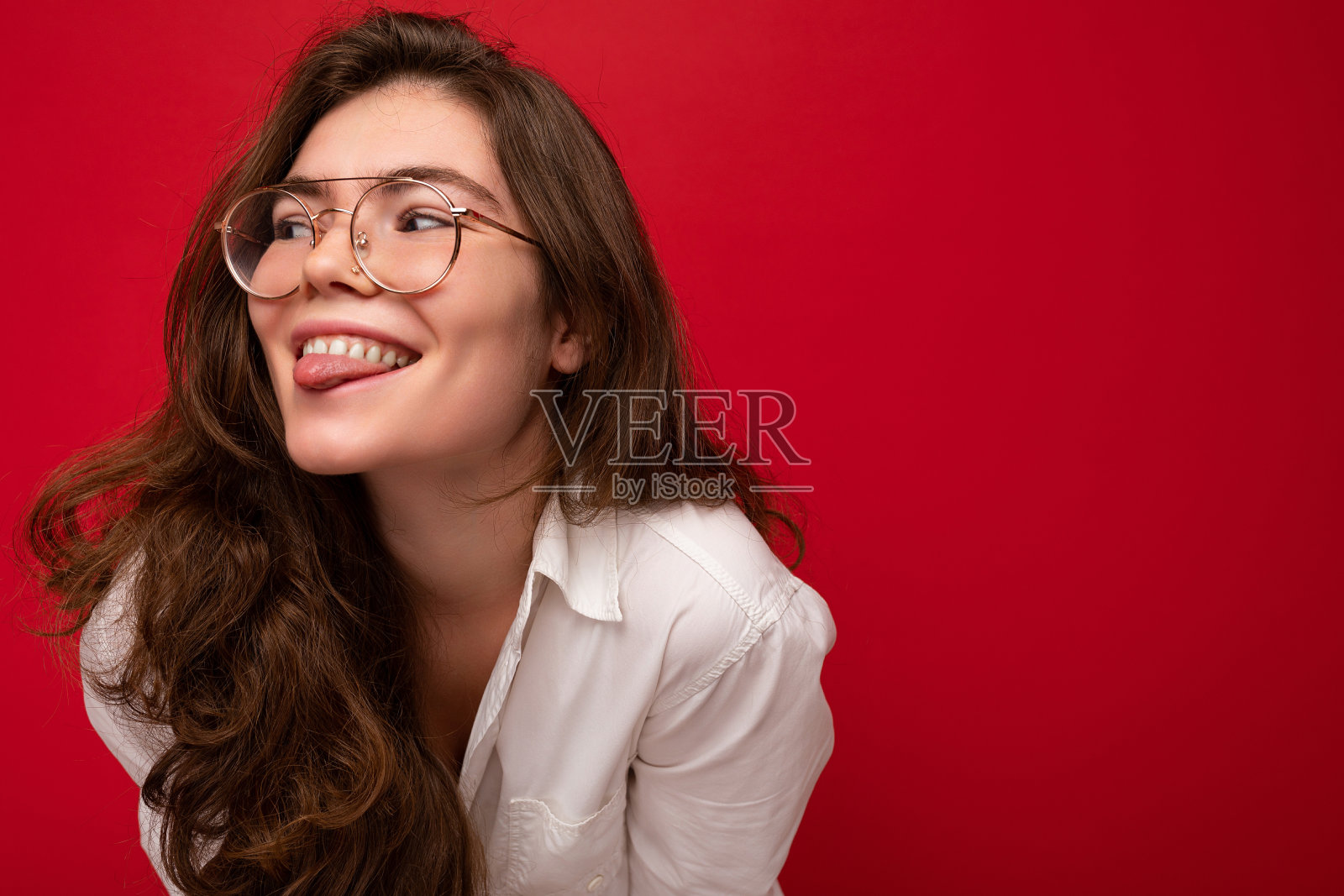 照片拍摄美丽性感快乐年轻的褐发卷女人孤立在红色背景穿着休闲的白色衬衫和时尚的眼镜看向一边和显示舌头照片摄影图片