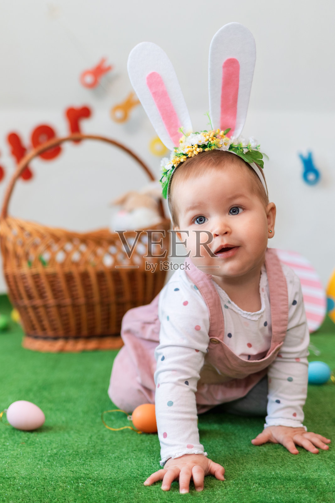 复活节。可爱的兔耳朵小女孩，在人造草坪上摆姿势。背景是一个篮子和鸡蛋。垂直照片摄影图片