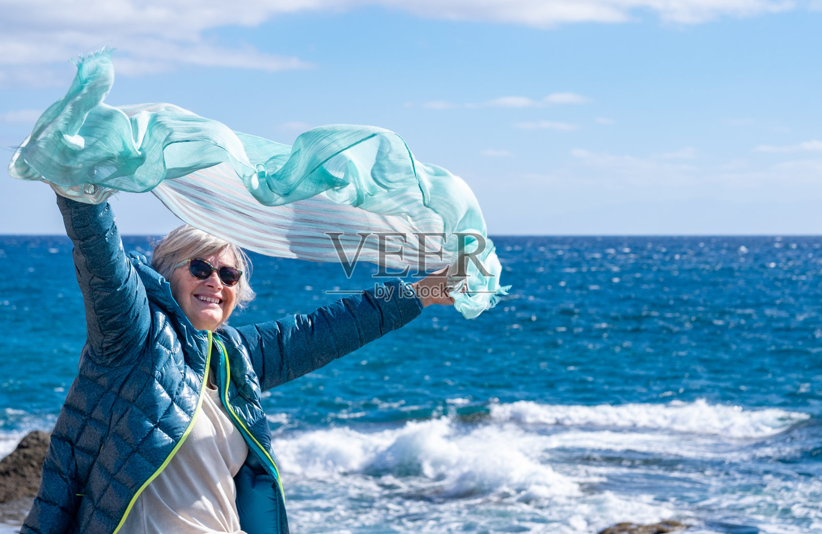 一个微笑放松的妇女面对大海的肖像，坐在悬崖上挥舞着她的围巾在风中。享受退休生活和户外生活的老年妇女照片摄影图片