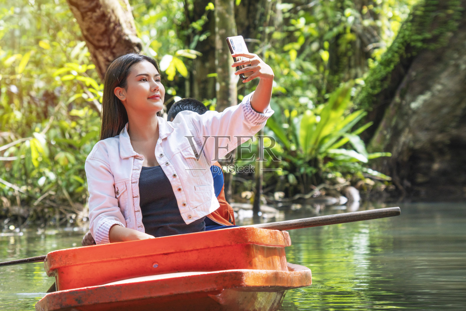 数字技术在假日的概念。享受年轻女子的长发使用智能手机自拍，而坐在船上休闲放松在湖与自然背景。照片摄影图片