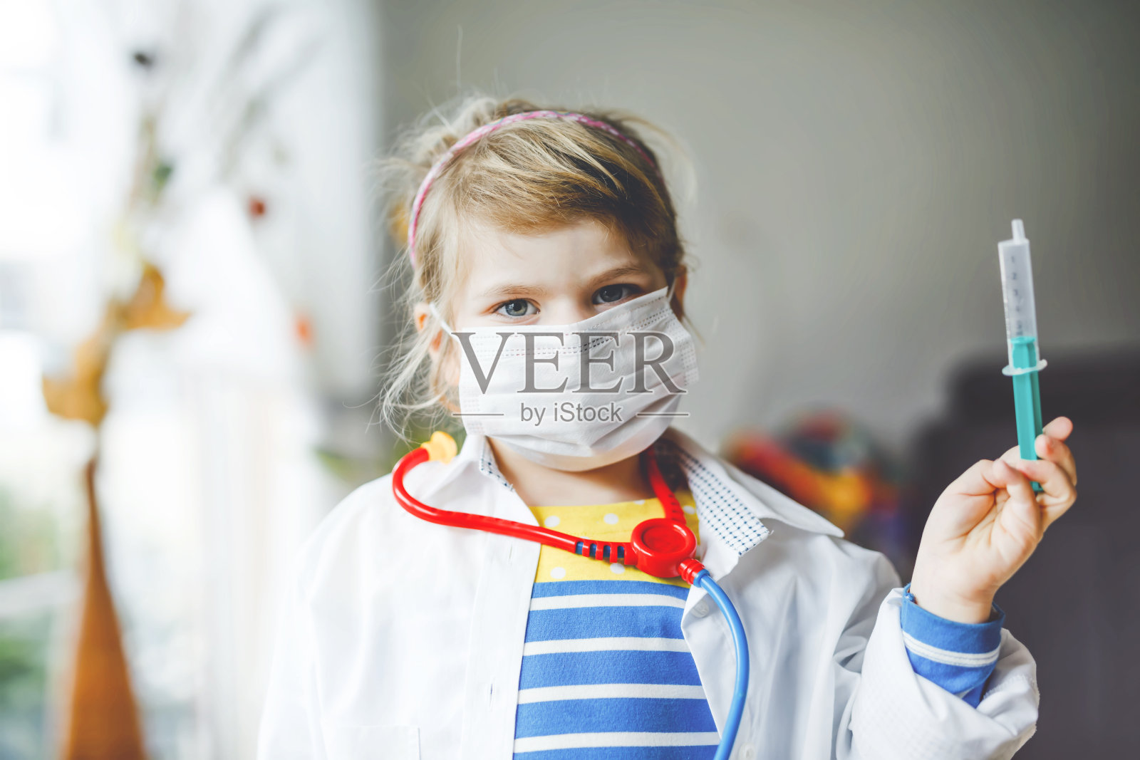 小女孩在打针。可爱的孩子戴着医用口罩扮演医生，拿着注射疫苗的注射器。冠状病毒疫苗概念。孩子在家里玩角色游戏。照片摄影图片