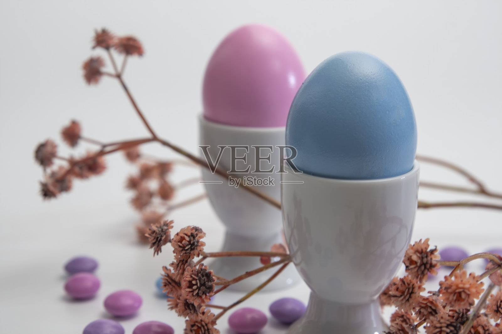 侧面的两个瓷质蛋杯与粉红色和蓝色的复活节蛋在白色的背景和小花。照片摄影图片