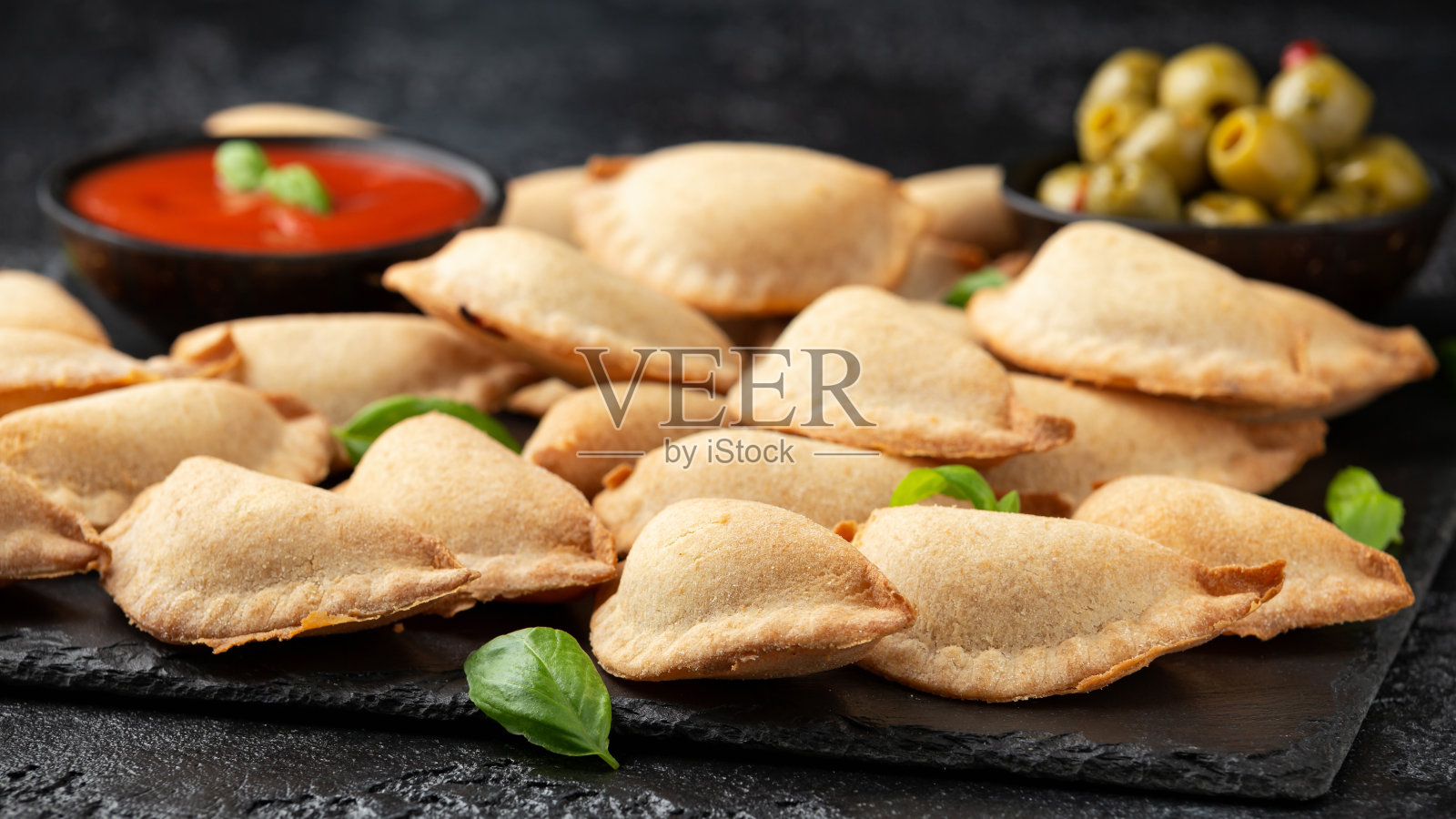 玛格丽塔披萨是一种包有西红柿、马苏里拉奶酪和罗勒的小袋披萨，配上番茄沙司和橄榄，放在黑色板板上。手指食物照片摄影图片