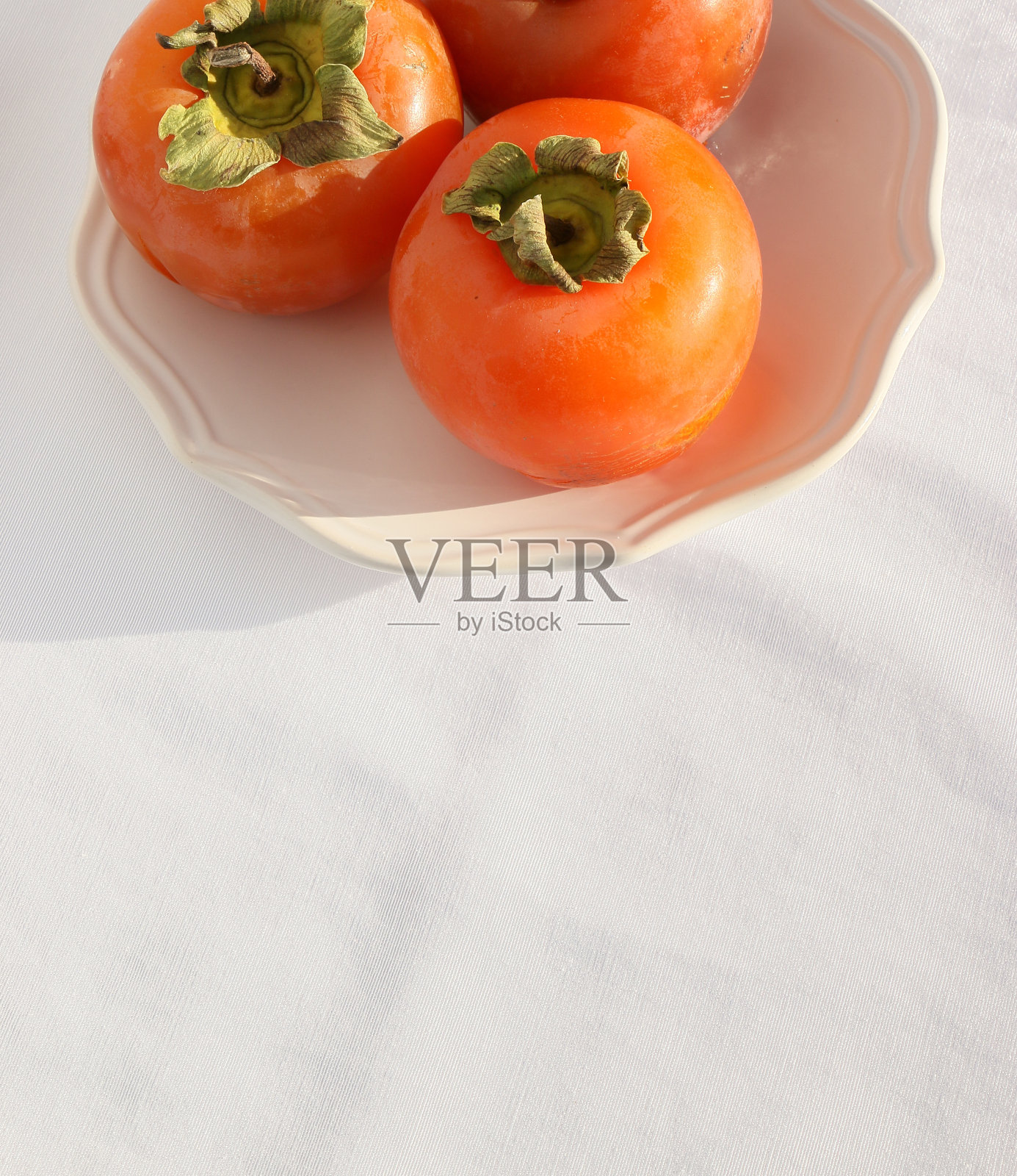 新鲜成熟的柿子放在白色盘子上，白色桌布上有经典的图案，这是棕榈天堂照片摄影图片