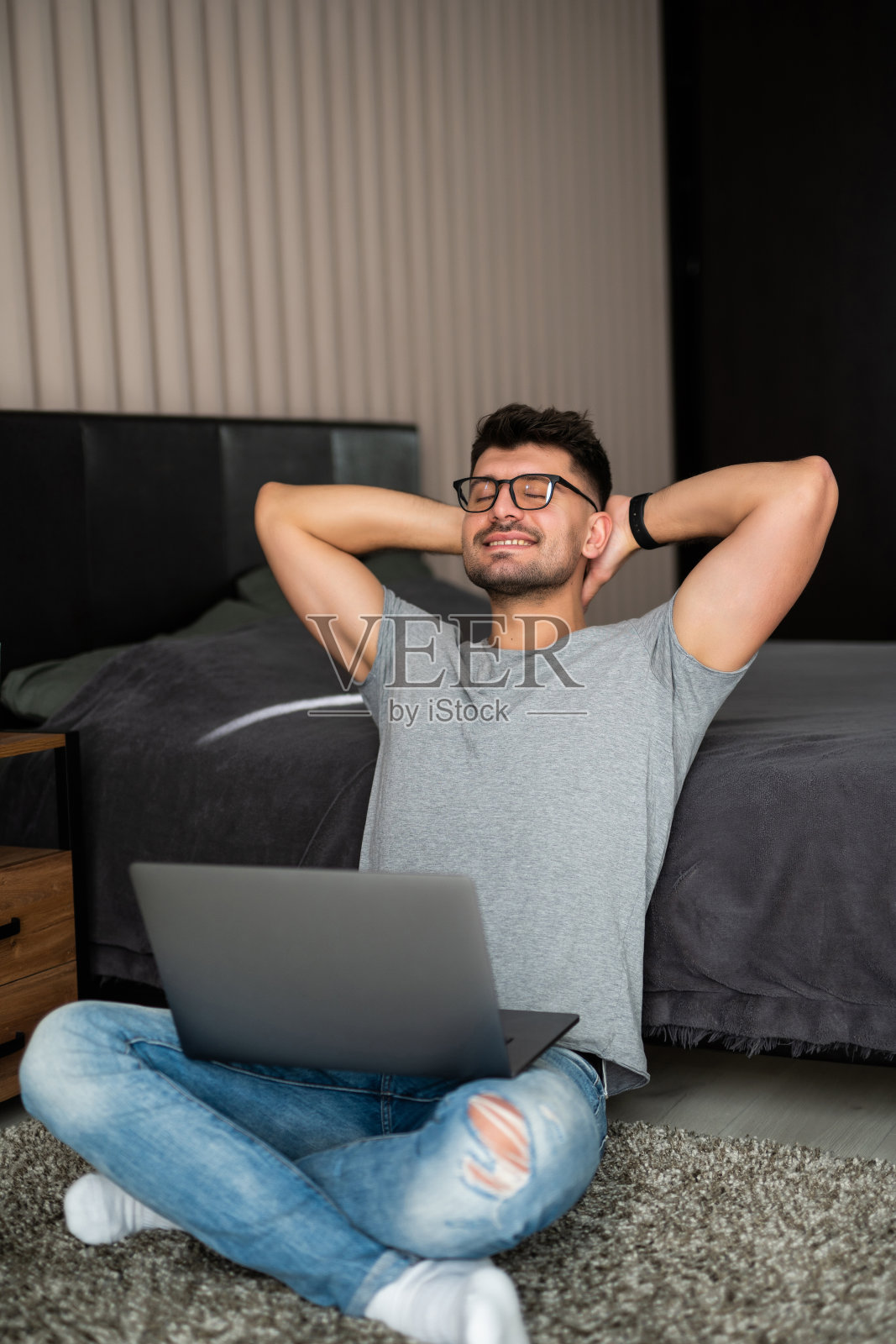 有魅力的大胡子男子坐在舒适的地毯地板上，穿着牛仔牛仔裤和休闲衬衫，在室内使用笔记本电脑照片摄影图片