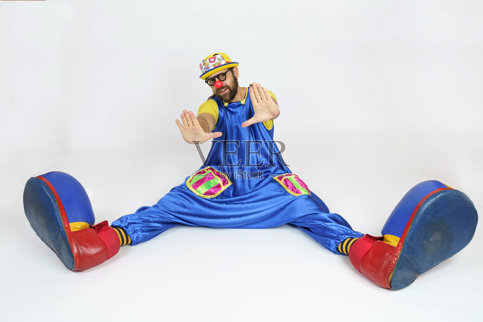 一个穿着鲜艳服装的小丑坐在那里，双腿分开，双手向前，手掌。照片摄影图片